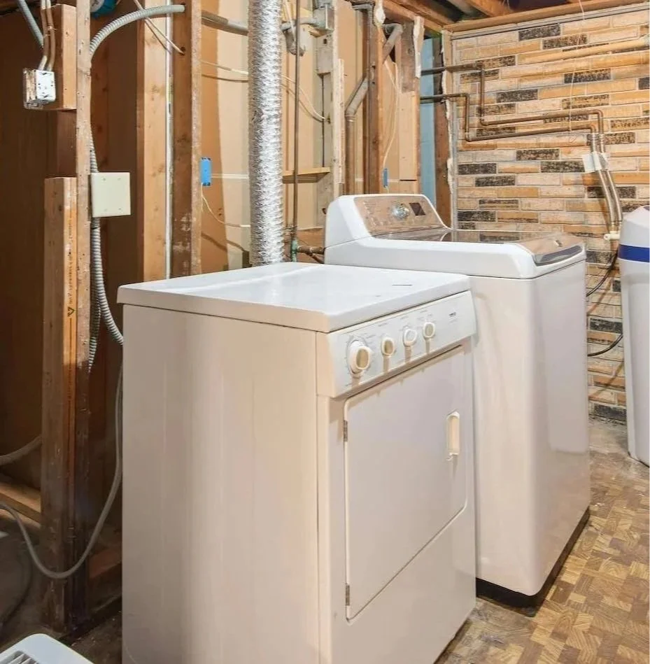 A laundry room with a white dryer, a white washing machine, and another appliance partially visible, all set against an unfinished wooden wall with exposed pipes and a brick accent wall.
