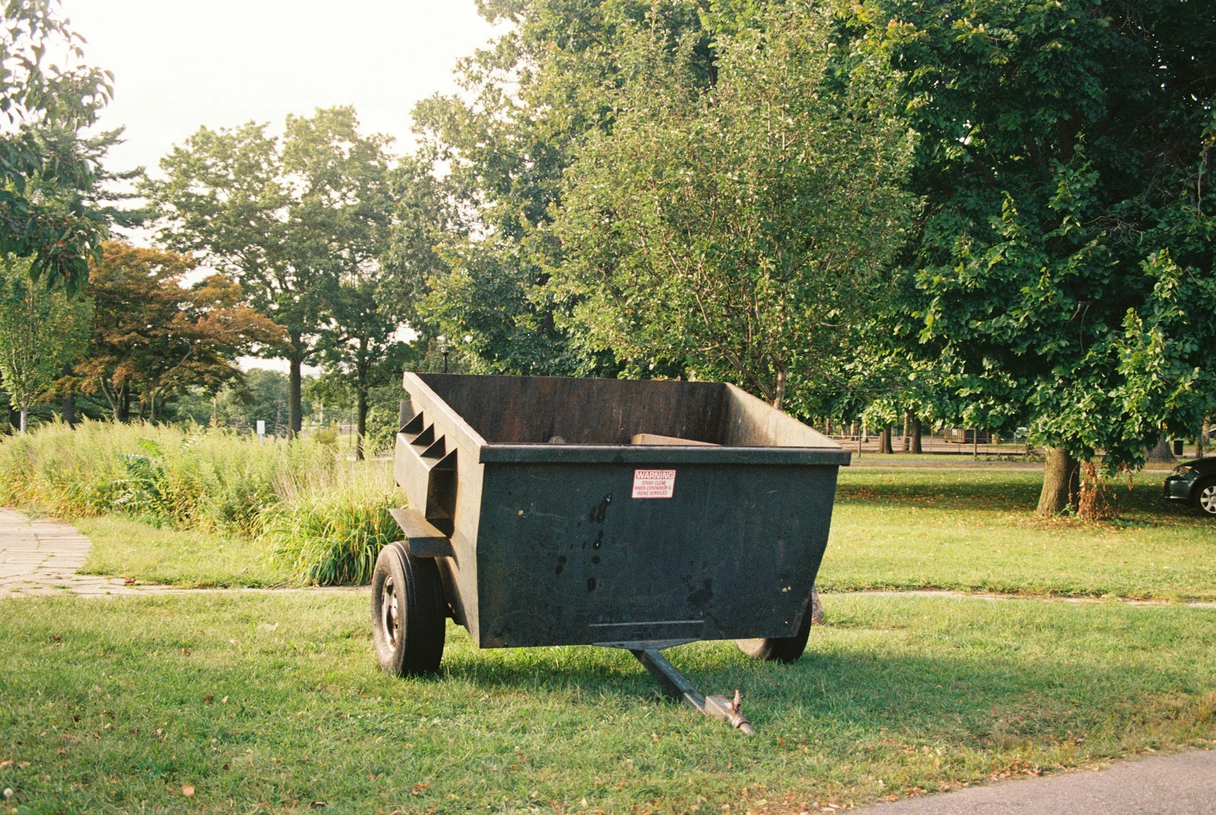 Black dumpster on a grassy area with trees in the background, some parked cars visible in the distance.