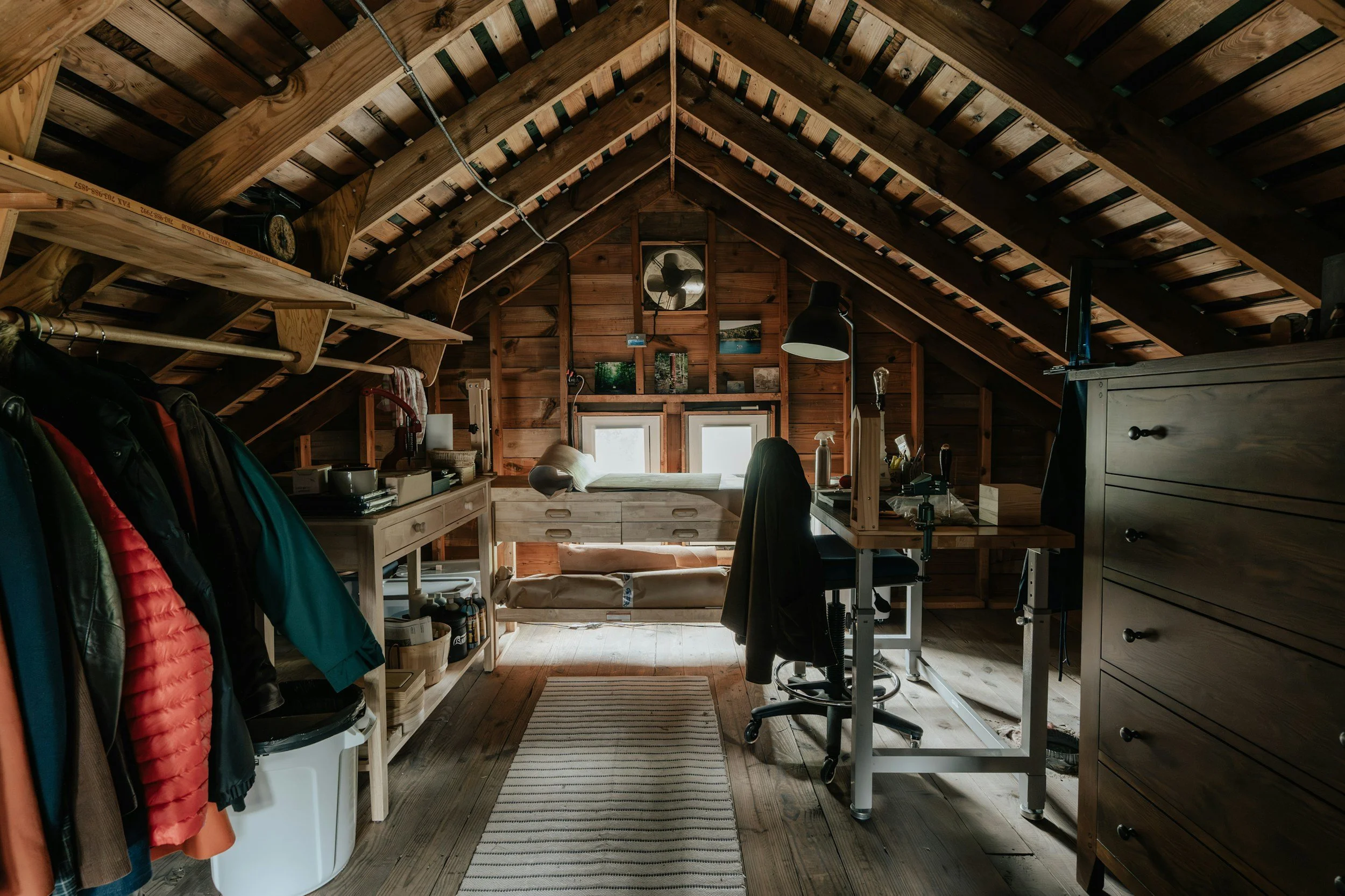 Attic workspace with wooden walls, ceiling, and floors, featuring a desk, chair, storage drawers, coats hanging, and windows at the far end.
