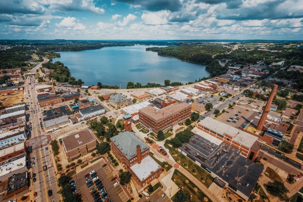 Aerial view of a small town with a large lake in the background, featuring old industrial buildings, streets, and parking lots.