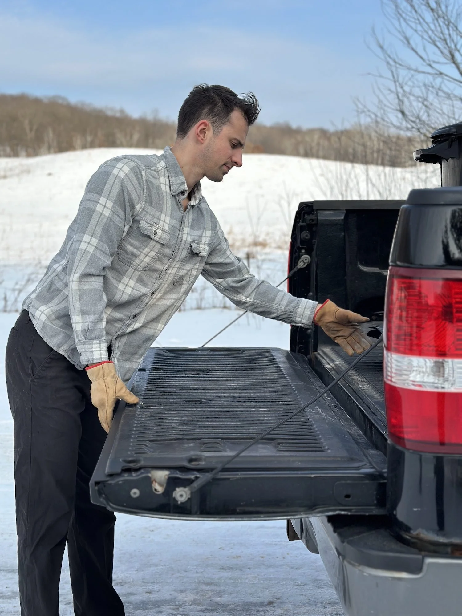 A man wearing a gray plaid shirt and tan gloves loading or unloading a black tailgate on a black pickup truck in a snowy outdoor setting.