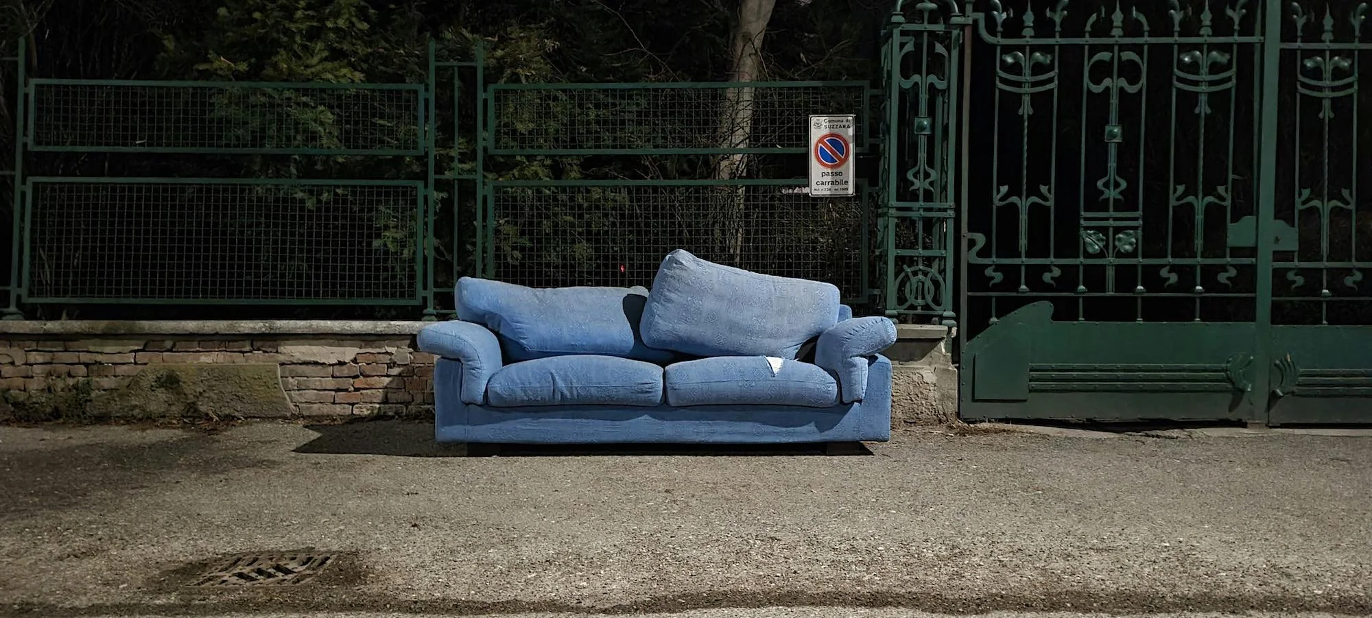 Blue couch placed on a sidewalk in front of a green metal gate and fencing at night.