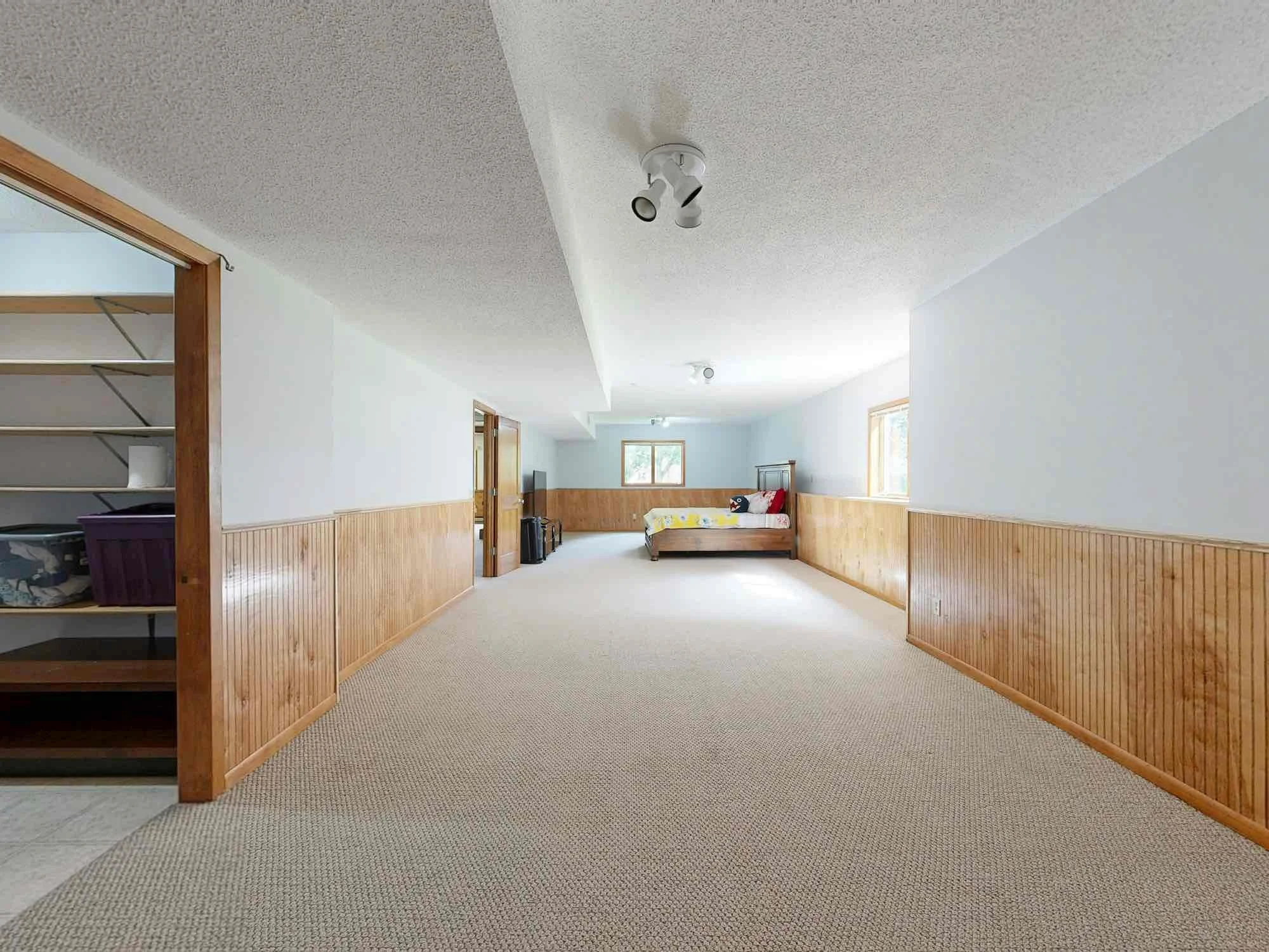 A spacious room with white walls and a carpeted floor, featuring wood paneling along the lower half of the walls. There are two windows letting in natural light, and on the far side, a bed with a colorful blanket and pillows. The ceiling has two light fixtures, and there are various items including a TV and some suitcases near the wall.