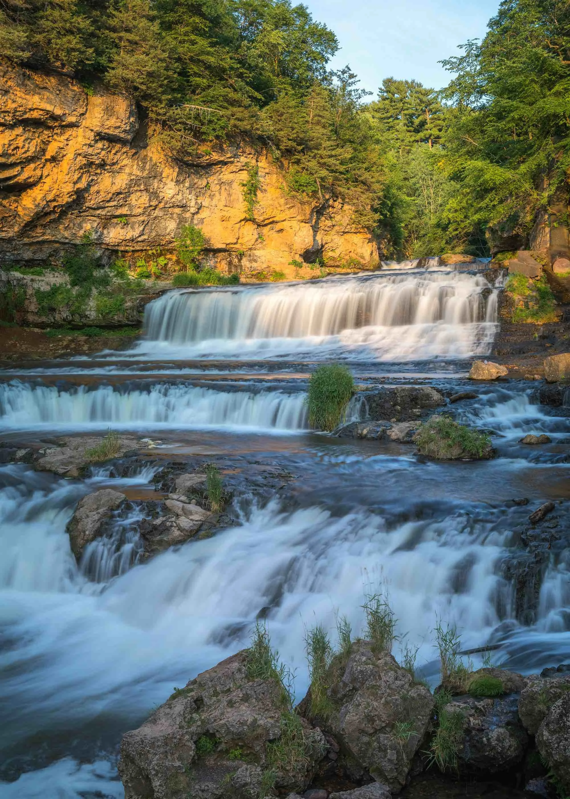 A multi-tiered waterfall flowing over rocks surrounded by trees and greenery in a forested area.