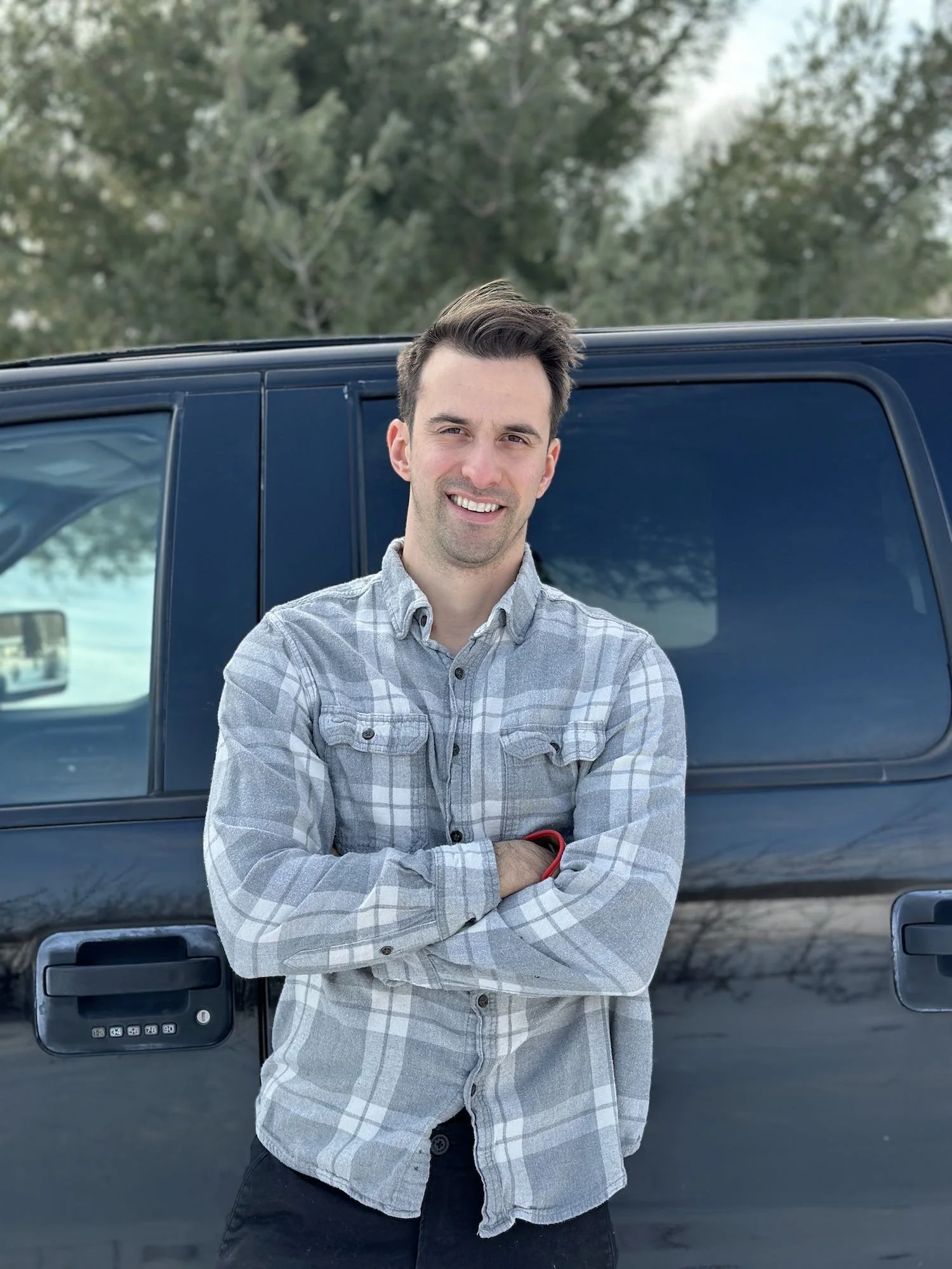 A young man with dark hair, wearing a gray plaid shirt, standing with arms crossed and smiling, in front of a dark-colored SUV outdoors with trees in the background.