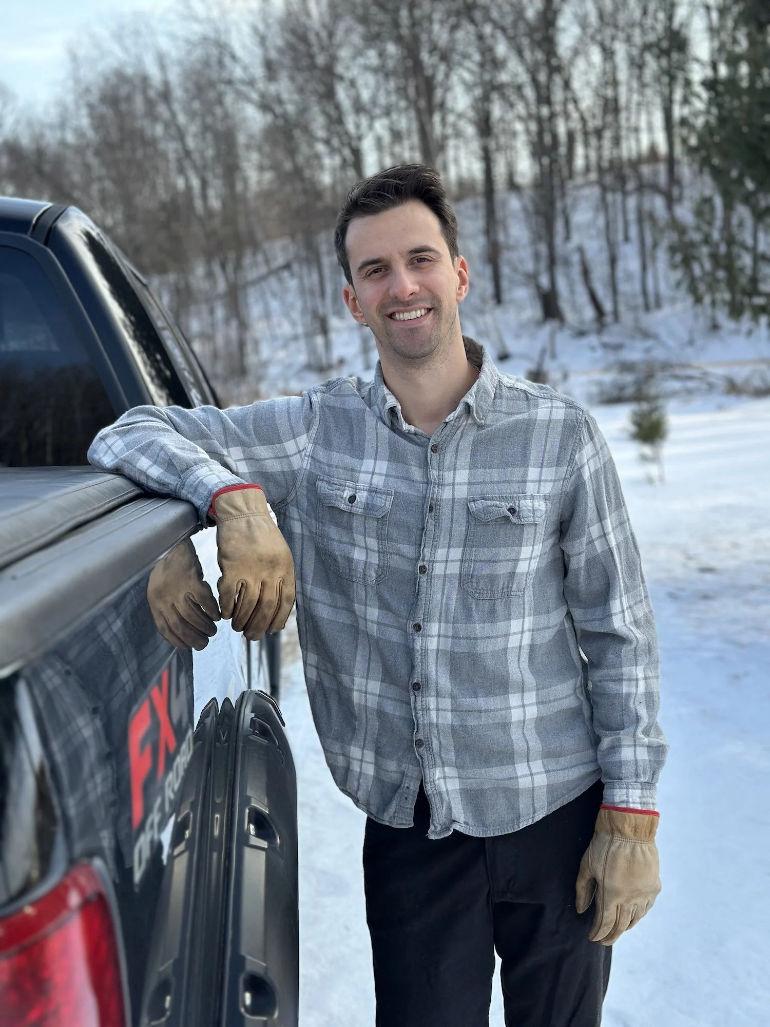 A young man with dark hair, smiling, wearing a gray plaid shirt and tan work gloves, leans on the side of a black truck with a snowy landscape and leafless trees in the background.