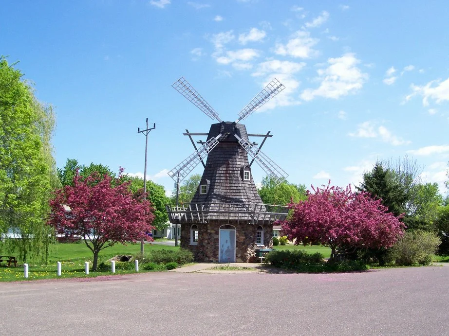 A small stone windmill with four blades, surrounded by pink flowering trees and green grass on a bright, sunny day.