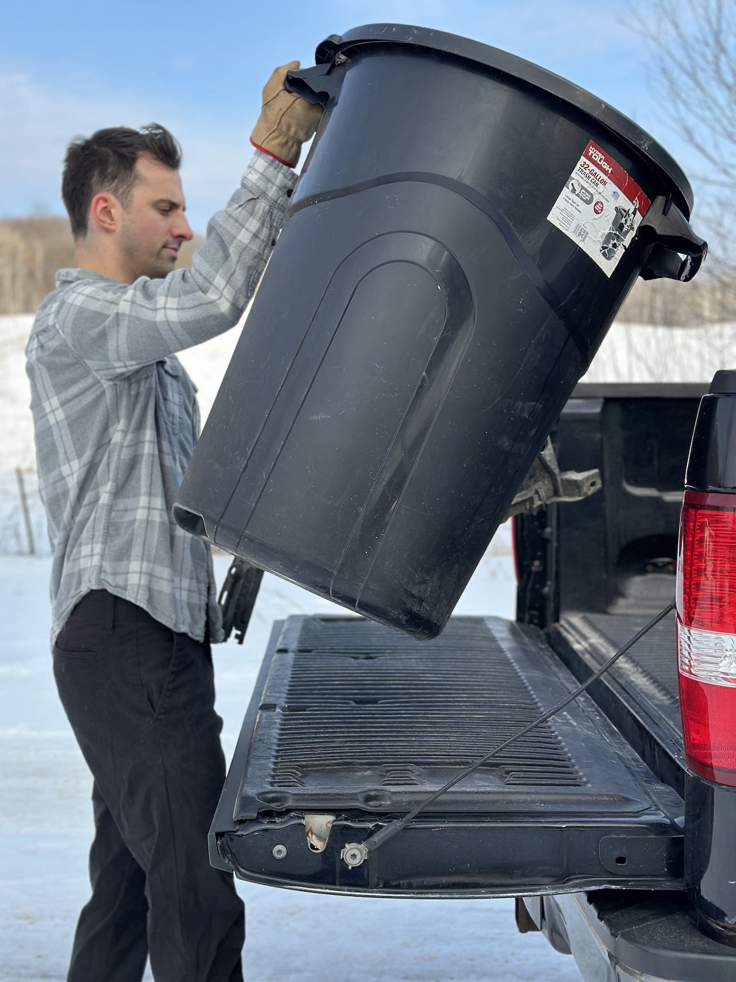 A man wearing a gray plaid shirt and gloves lifting a large black trash can from the back of a pickup truck in a snowy outdoor setting.