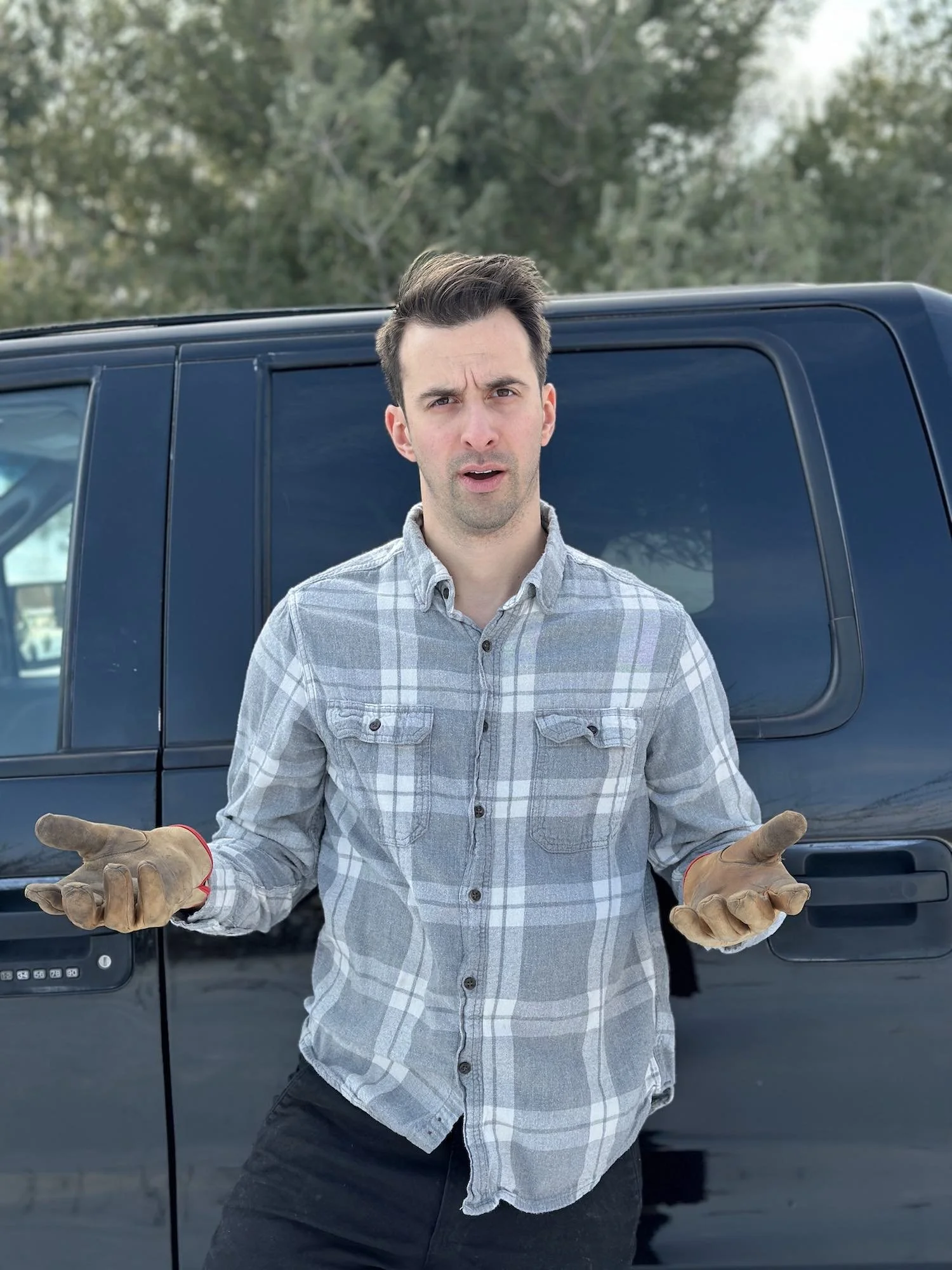 A young man with short dark hair and light skin wearing a gray plaid shirt and brown work gloves standing in front of a dark blue vehicle outdoors with leafy trees in the background, expressing confusion or questioning.