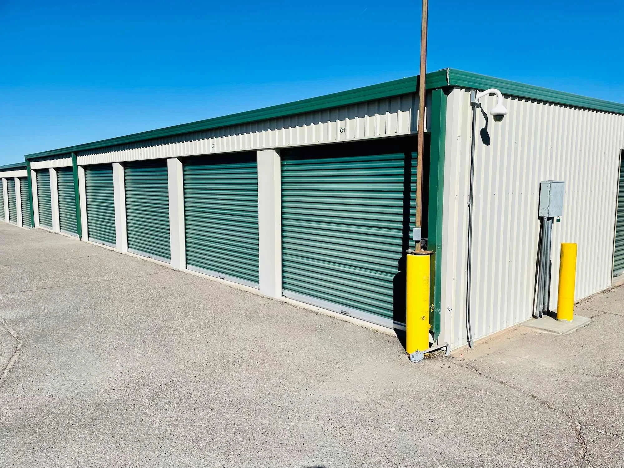 Row of storage units with green roll-up doors, white and green metal siding, and a security light on the corner; blue sky overhead.