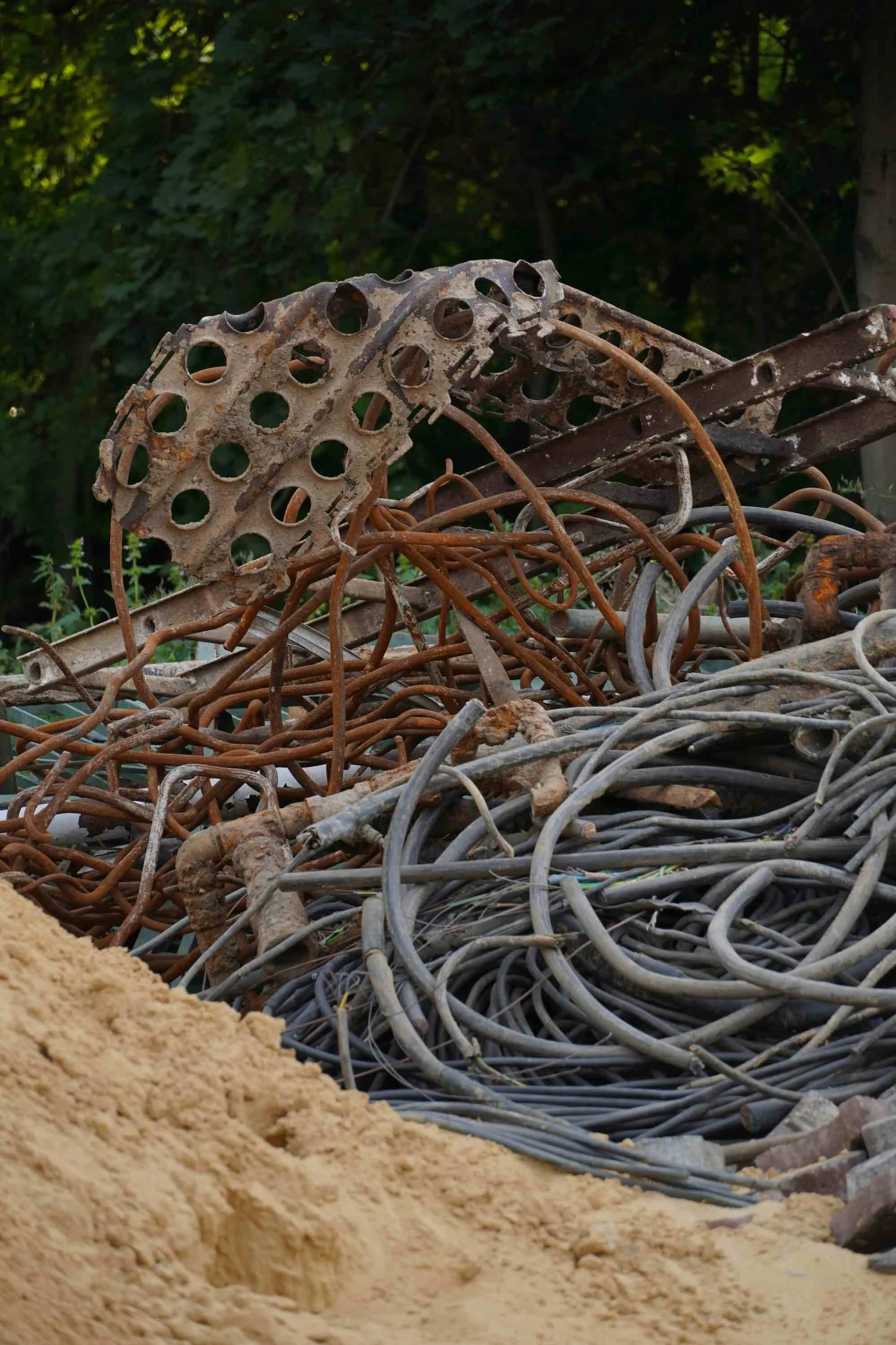 Pile of rusty metal scrap and wires with a sandy foreground and green trees in the background.