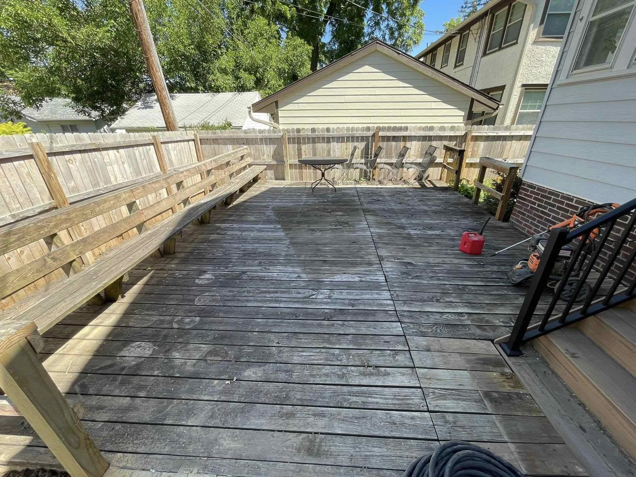 Backyard wooden deck with a long bench along the left side, a small table and several chairs at the back, a red gas can, and a string trimmer near the house on the right. Fencing encloses the yard, with neighboring houses visible beyond.