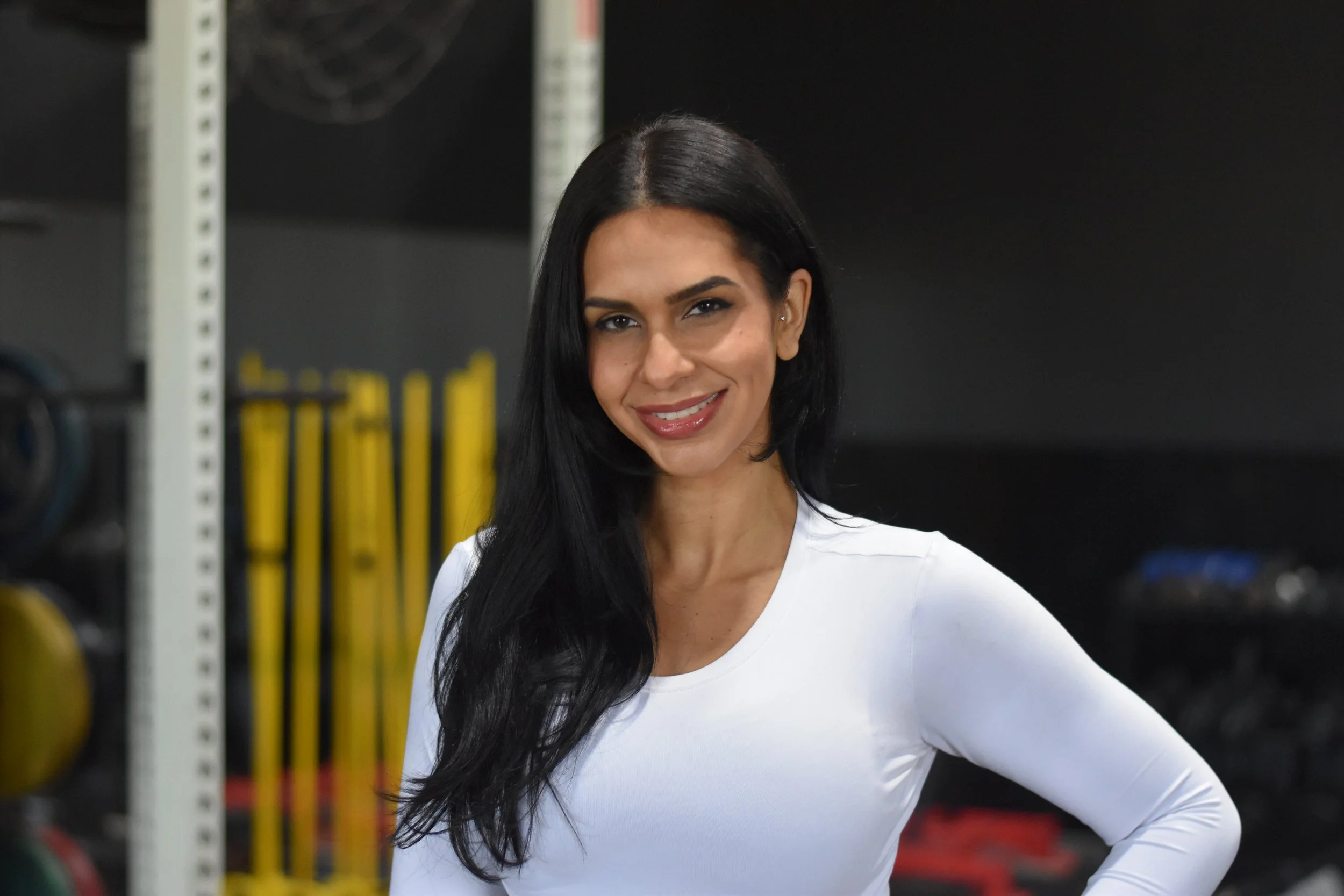 Smiling woman with long black hair wearing a white shirt in a gym setting.