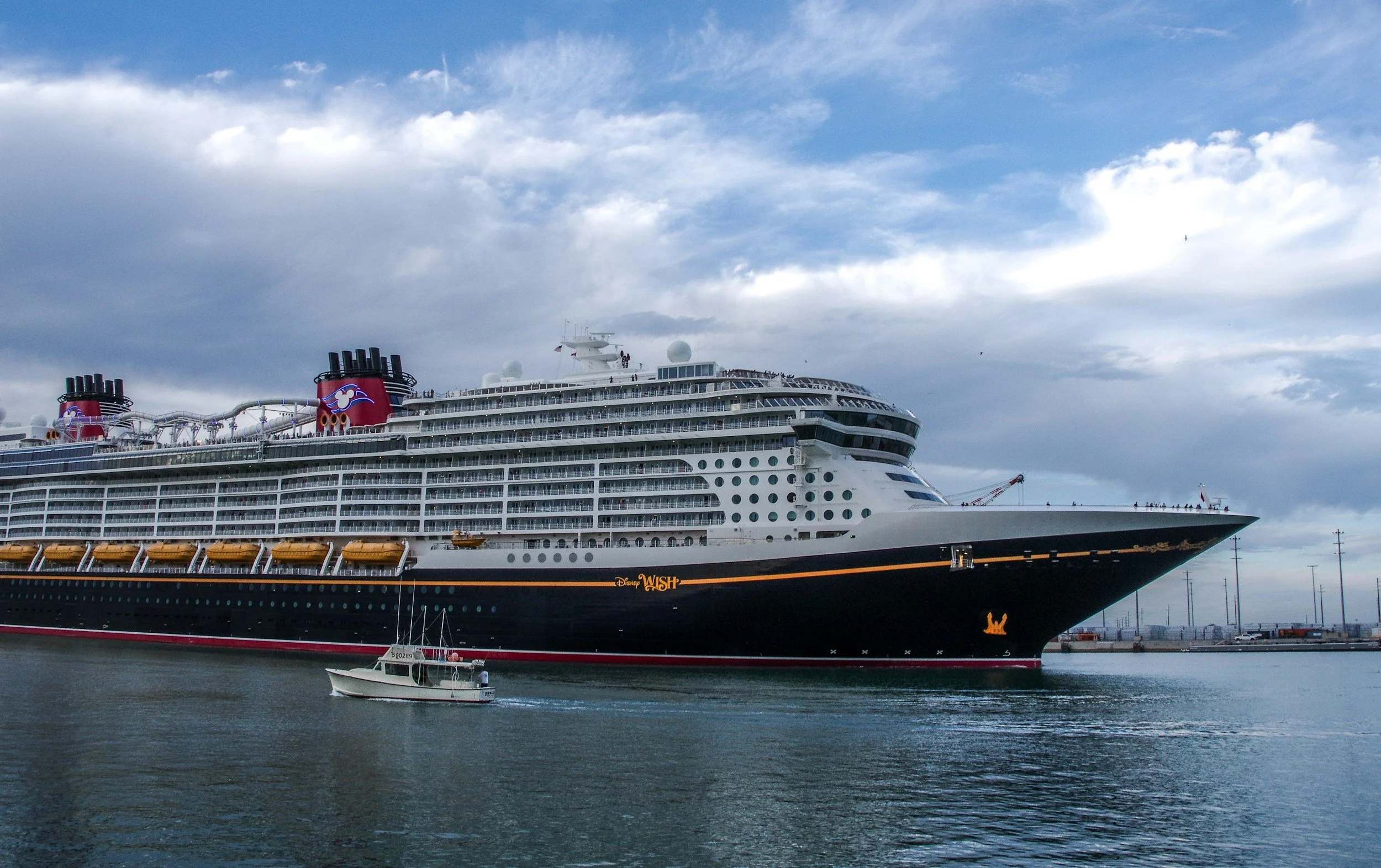 Large cruise ship named Disney Wish sailing in water, with a smaller boat nearby and a cloudy sky overhead.