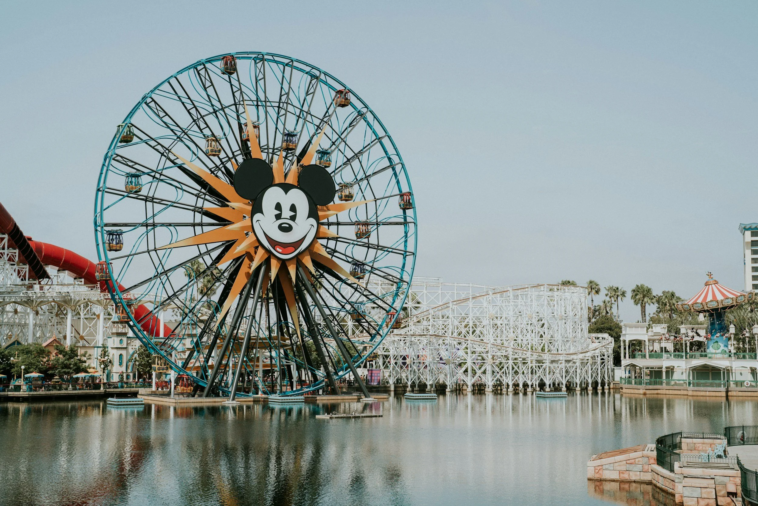 A large ferris wheel with a Mickey Mouse face in the center, on a lakeside at an amusement park. The ride is blue and orange, with gondolas around the wheel. The park has a white wooden roller coaster and a carousel in the background, with palm trees and a clear sky.