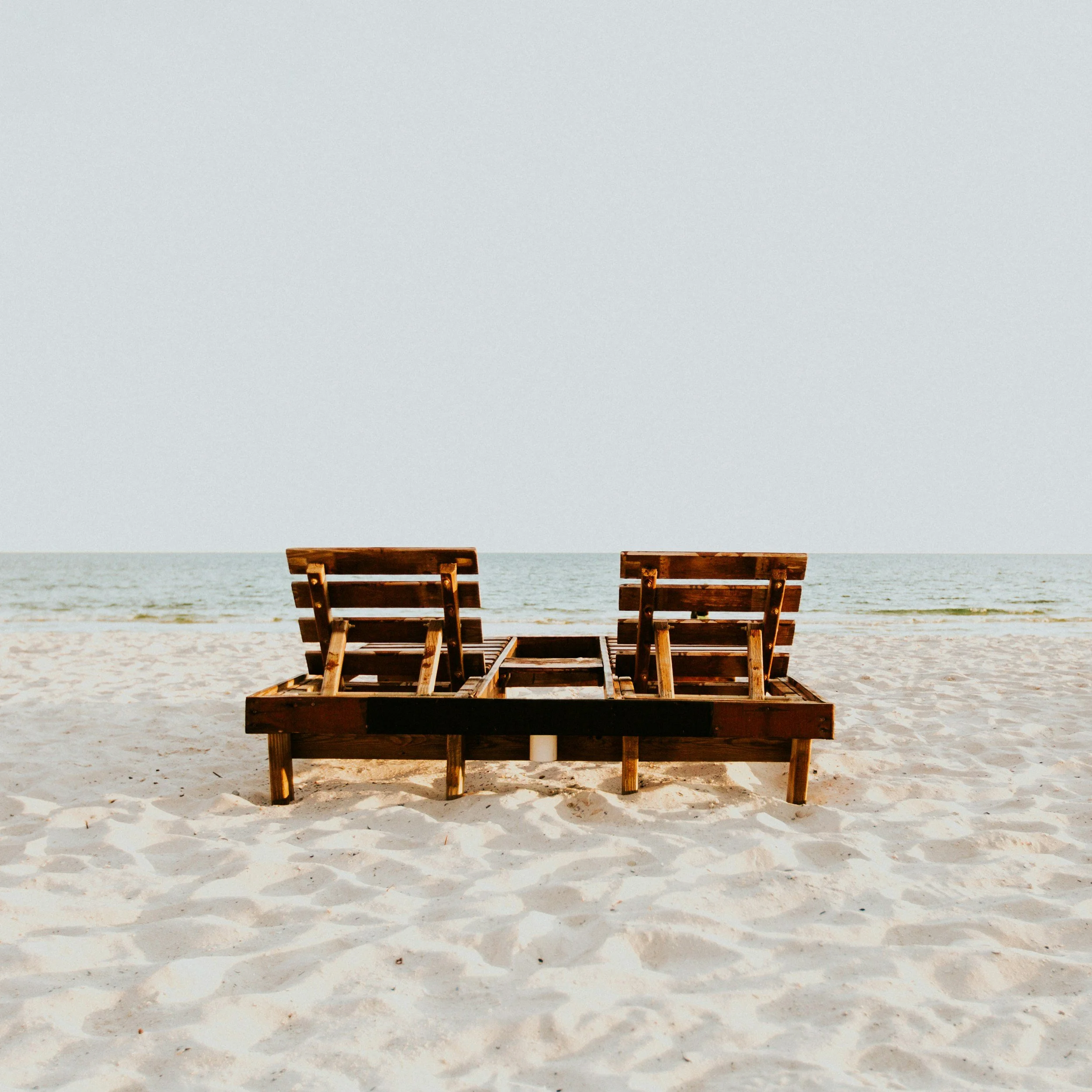 Two wooden lounge chairs on a sandy beach facing the ocean.