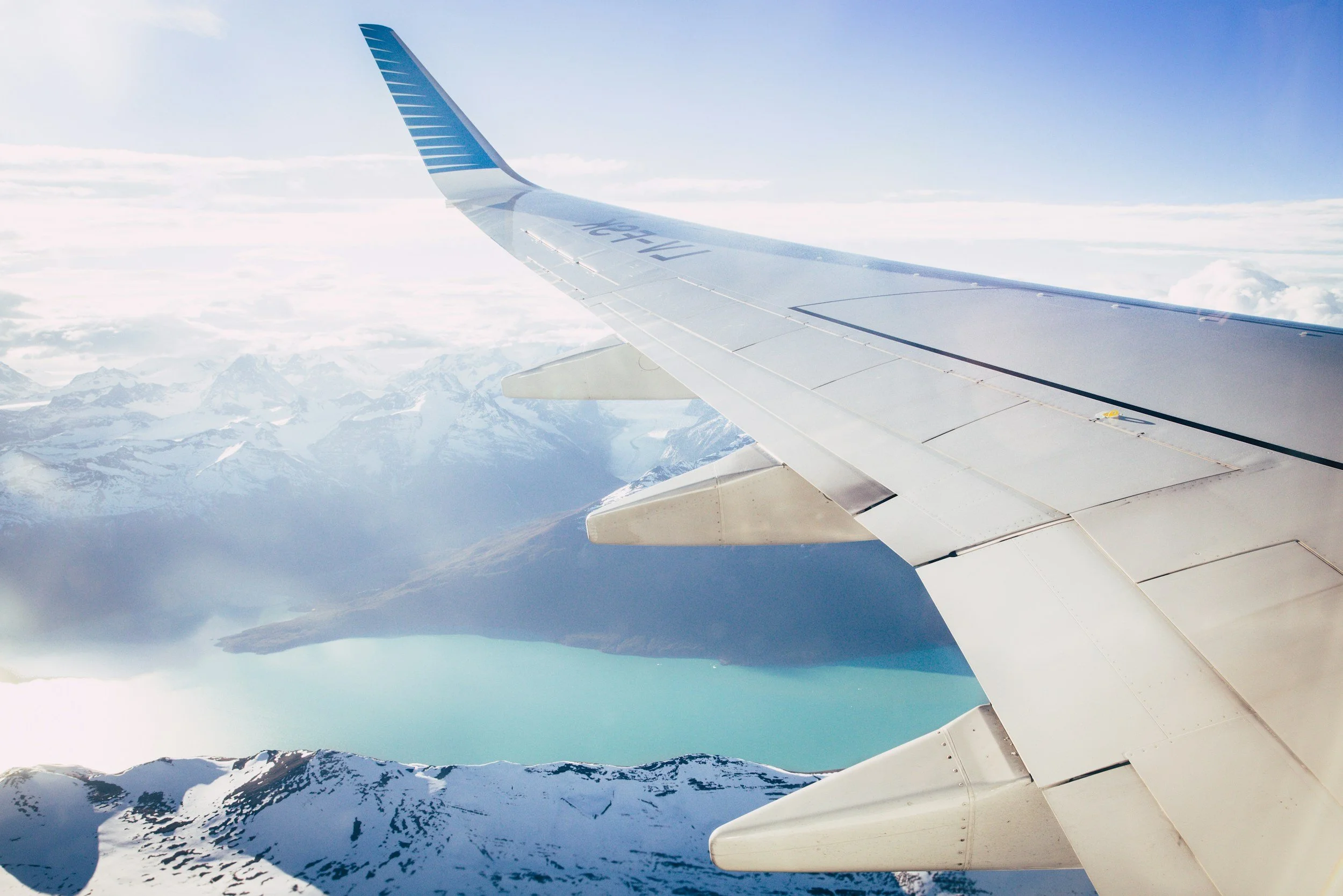 View from an airplane window showing the wing with snow-covered mountains and a lake below under a partly cloudy sky.