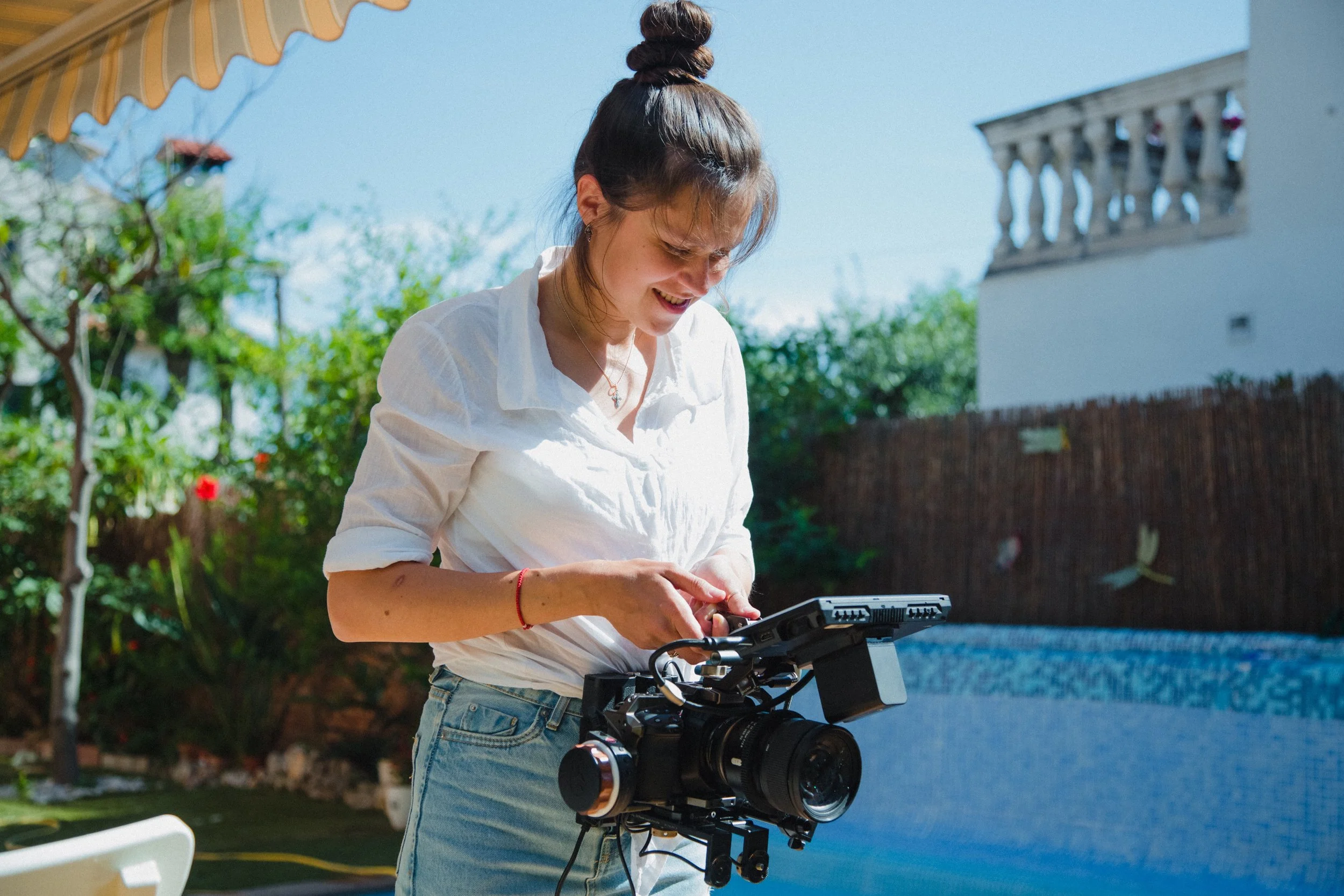 A woman with dark hair in a bun, wearing a white shirt, is operating a professional camera mounted on a tripod outdoors under a yellow and white striped awning. Trees and a building are visible in the background on a sunny day.