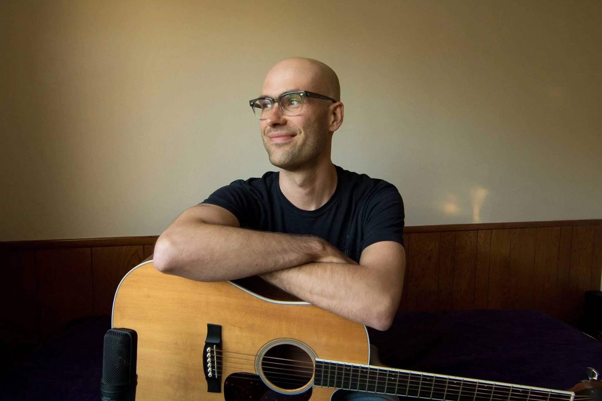 A man with glasses and a shaved head sitting at a table with a guitar, looking to the side, in a room with cream-colored walls and wooden paneling.