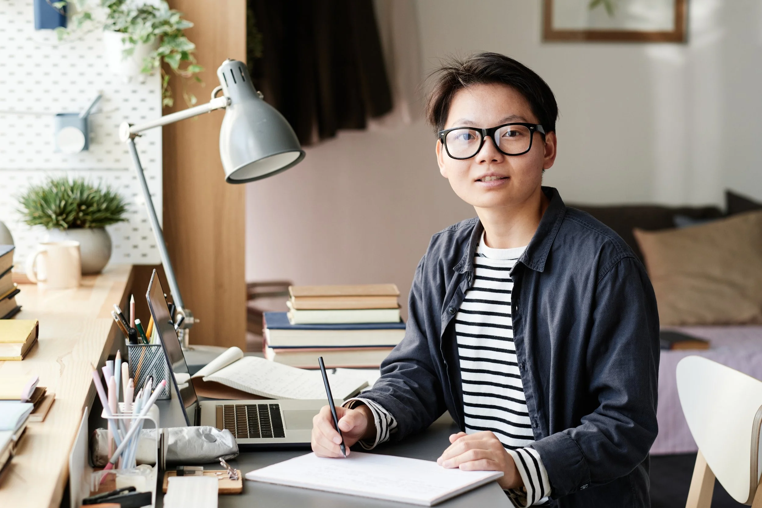 A young person with short dark hair, glasses, and a striped shirt, sitting at a cluttered desk, writing on paper with a pen, surrounded by books, a laptop, and various stationery items in a cozy home office.