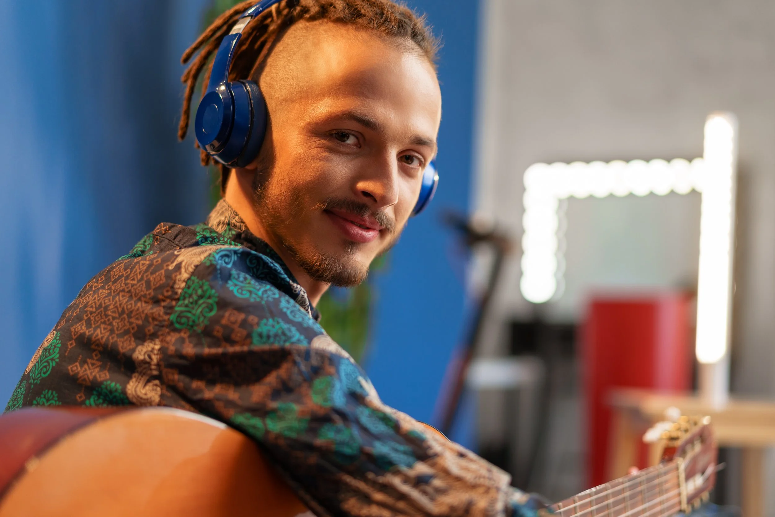 A man with a beard and dreadlocks wearing nature-inspired clothing, smiling while playing an acoustic guitar in a colorful room with blue walls and bright lighting.