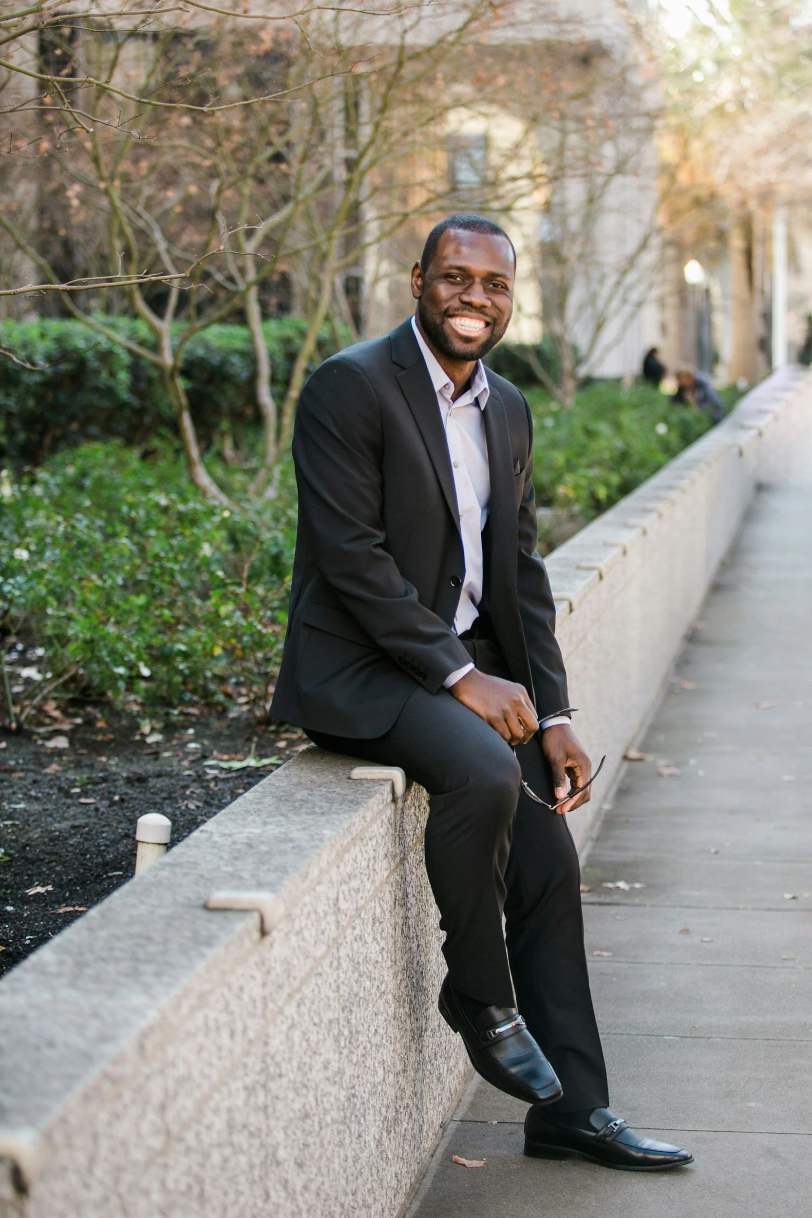 A man in a black suit sitting on a stone ledge outdoors, smiling, with trees and a building in the background.