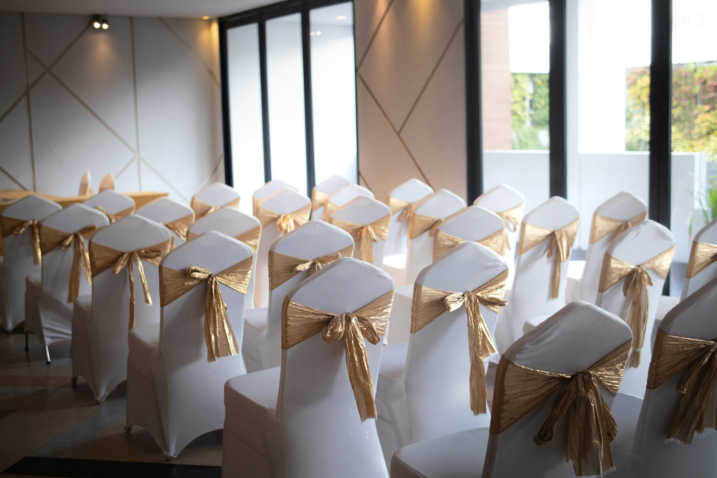 Rows of chairs decorated with white covers and gold bows, set up for a formal event in a room with large windows allowing natural light.
