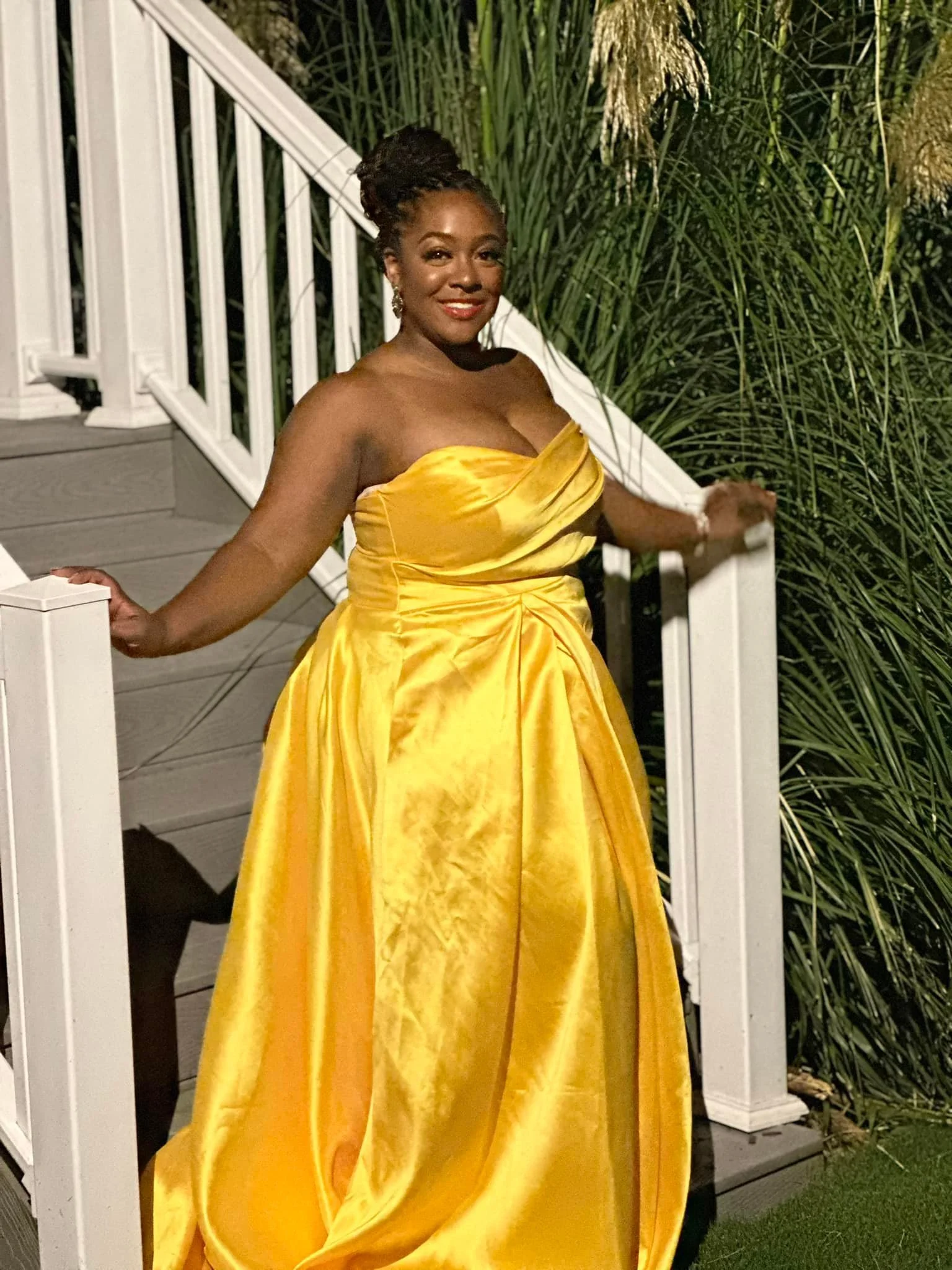 A woman in a strapless, bright yellow gown with a full skirt, standing outdoors at night near a white railing and lush green plants.
