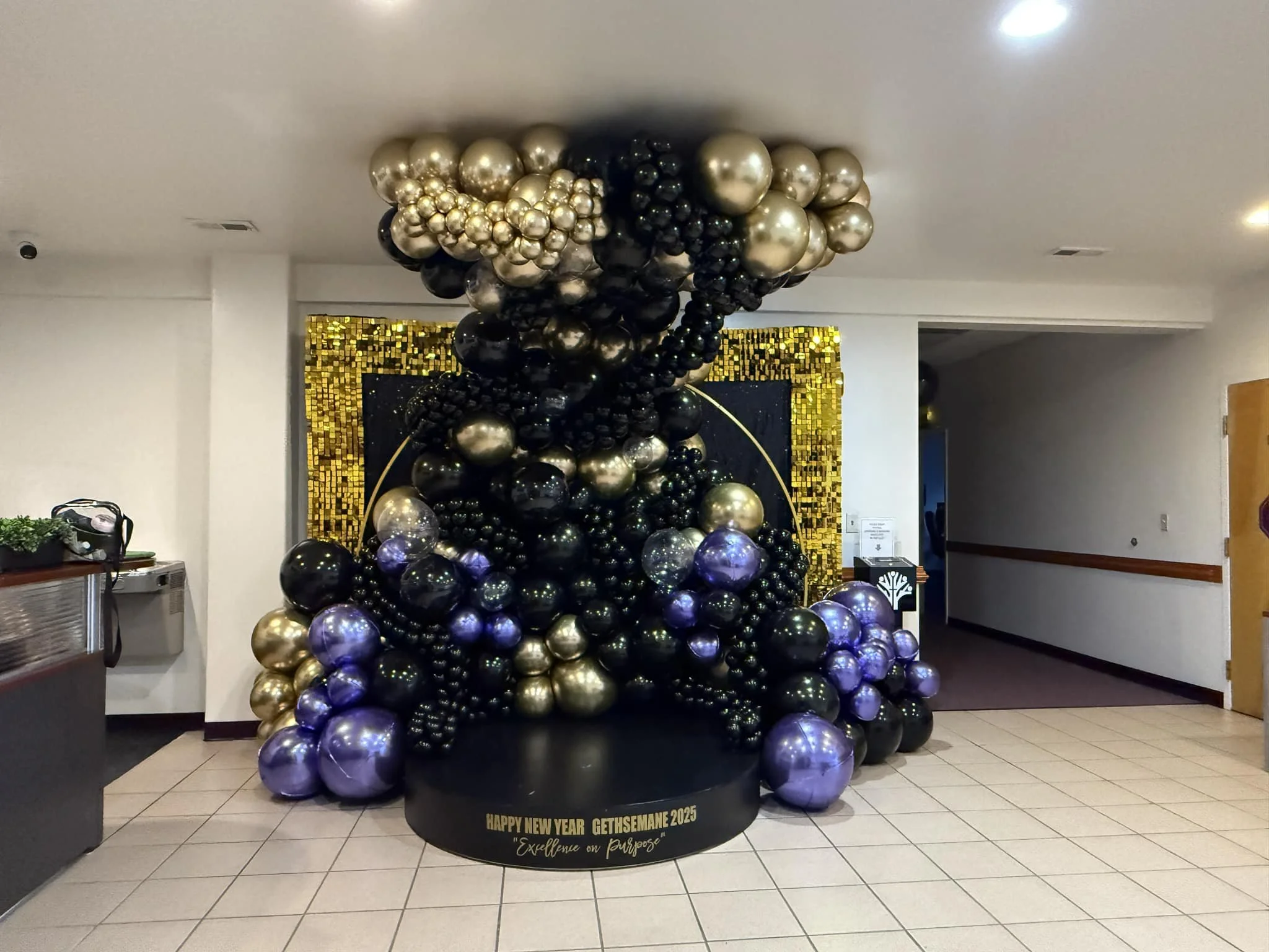 A decorative balloon arrangement in a hallway, featuring black, gold, and silver balloons, with a gold mosaic wall in the background. The arrangement is on a black base with a gold inscription reading "HAPPY NEW YEAR GETHSEMANE 2025 - Excellence on Purpose."