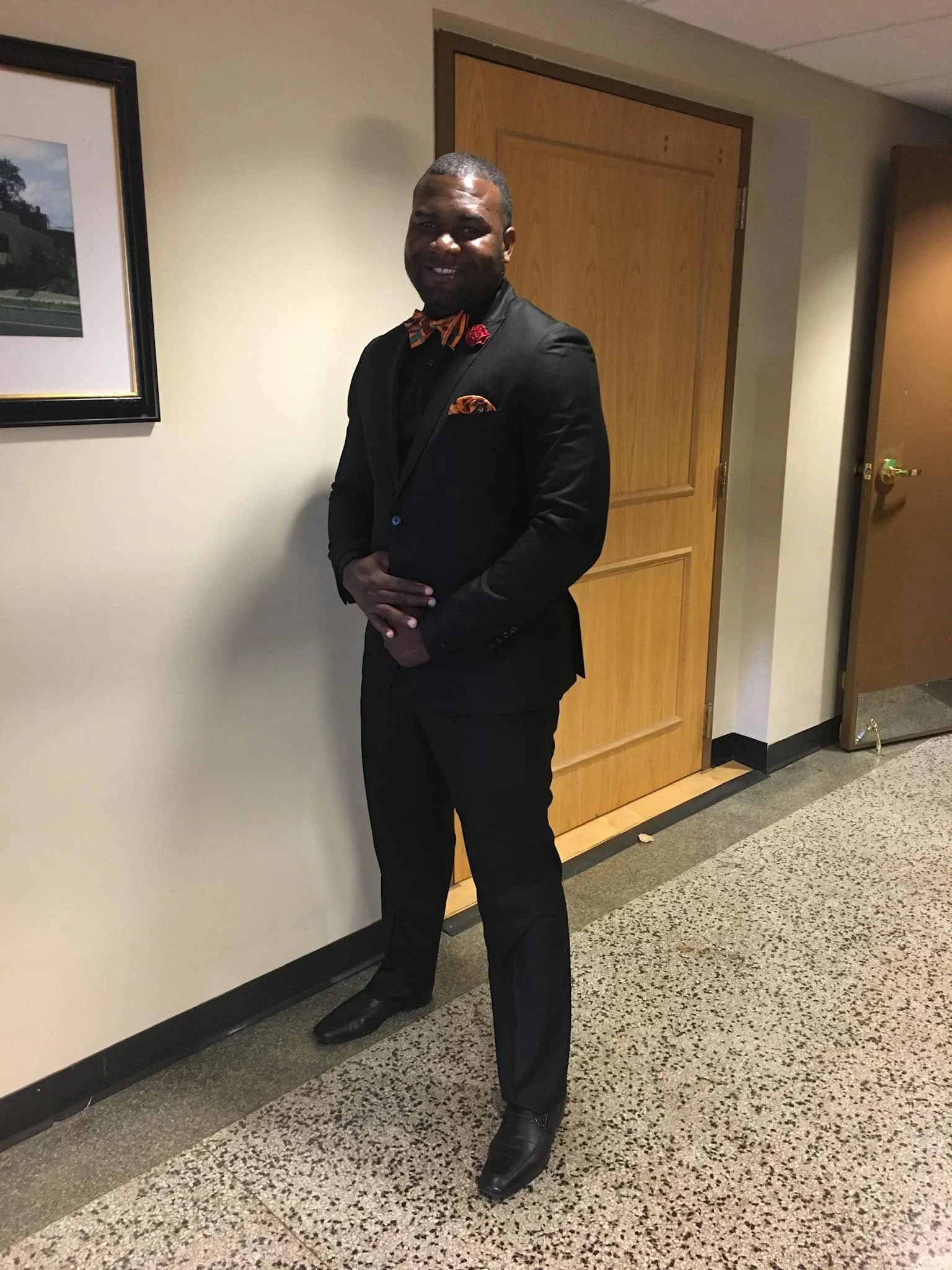 A man dressed in a formal black suit with colorful accessories, standing indoors against a wall near a wooden door, smiling at the camera.
