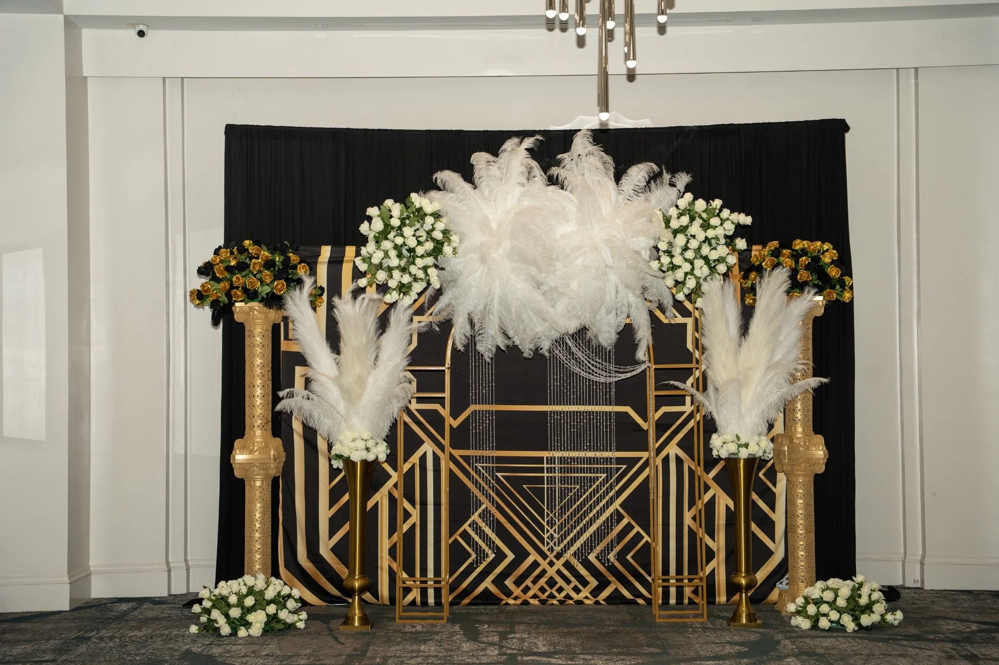Elegant black and gold geometric backdrop decorated with white feathers, gold and white roses, and white flower arrangements on tall gold vases, likely for a special event or celebration.