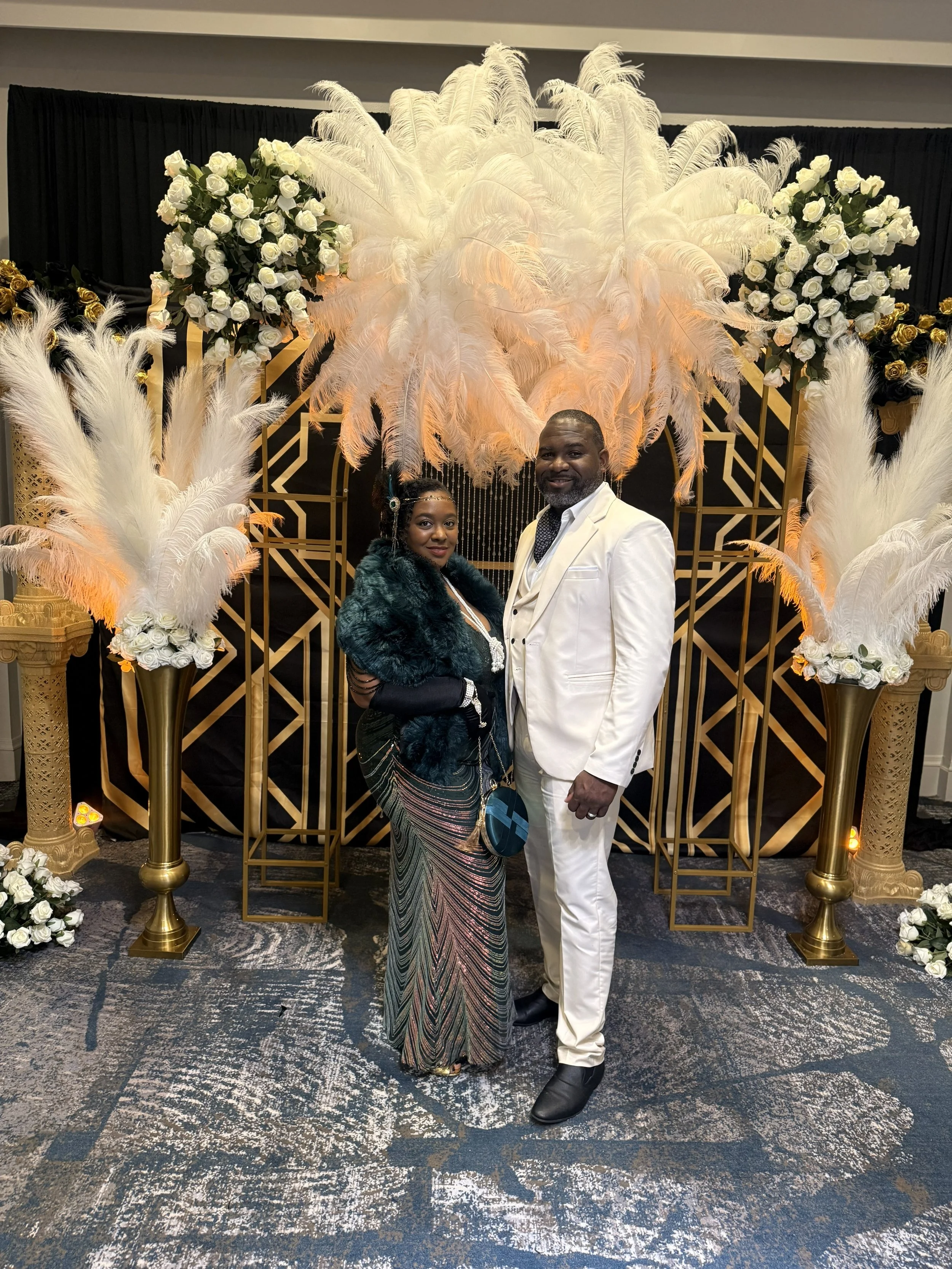 A couple standing in front of an elegant black, gold, and white decorative backdrop with feathers and roses, dressed in formal attire, at a special event or celebration.