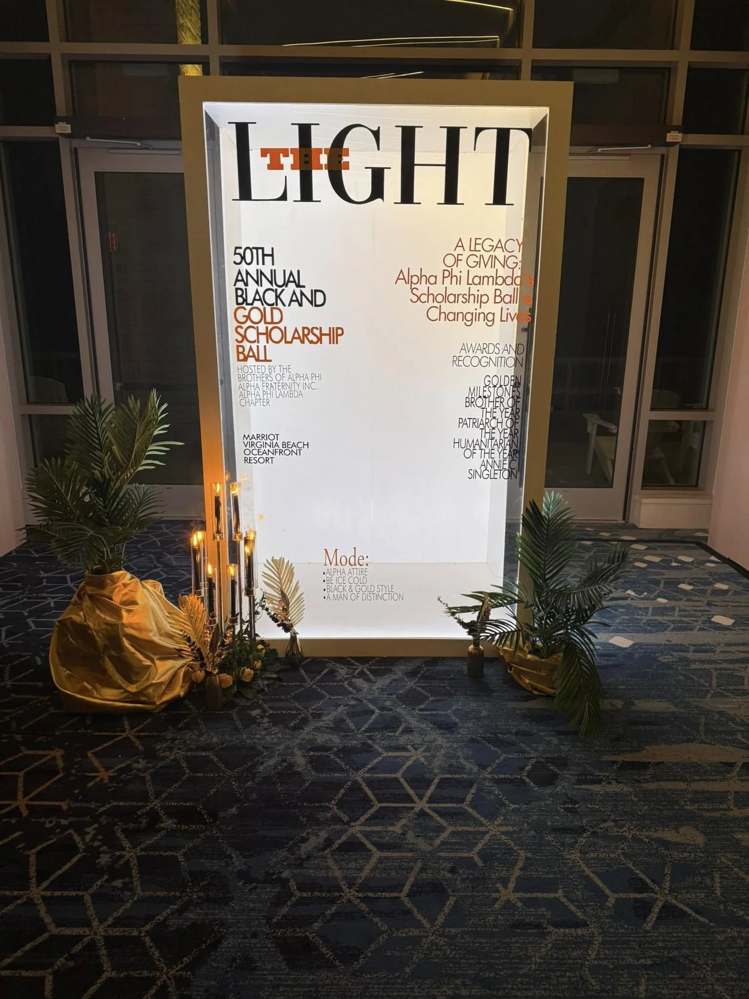 A large white illuminated sign with black and red text for an event called 'The Light.' The event is the 50th annual Black and Gold Scholarship Ball, hosted by the Brothers of Alpha Phi Alpha Fraternity Inc. The sign lists highlights such as awards and recognition, gold milestones, and the Patriot of the Year. Flowers and greenery decorate the bottom of the sign, and a patterned carpet is visible on the floor.
