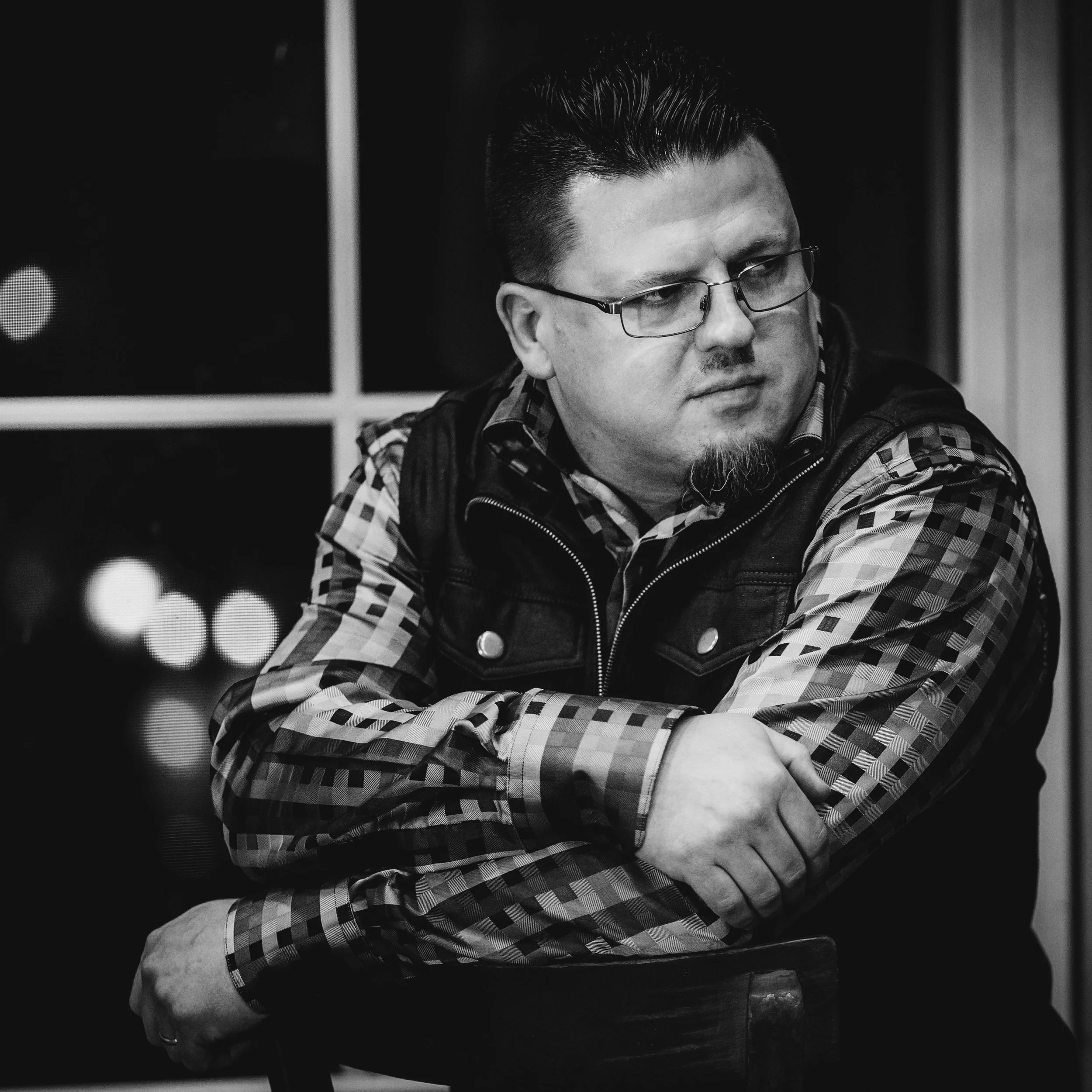 A black-and-white photo of a man with glasses, short dark hair, and a goatee, sitting with his arms crossed, looking out a window.