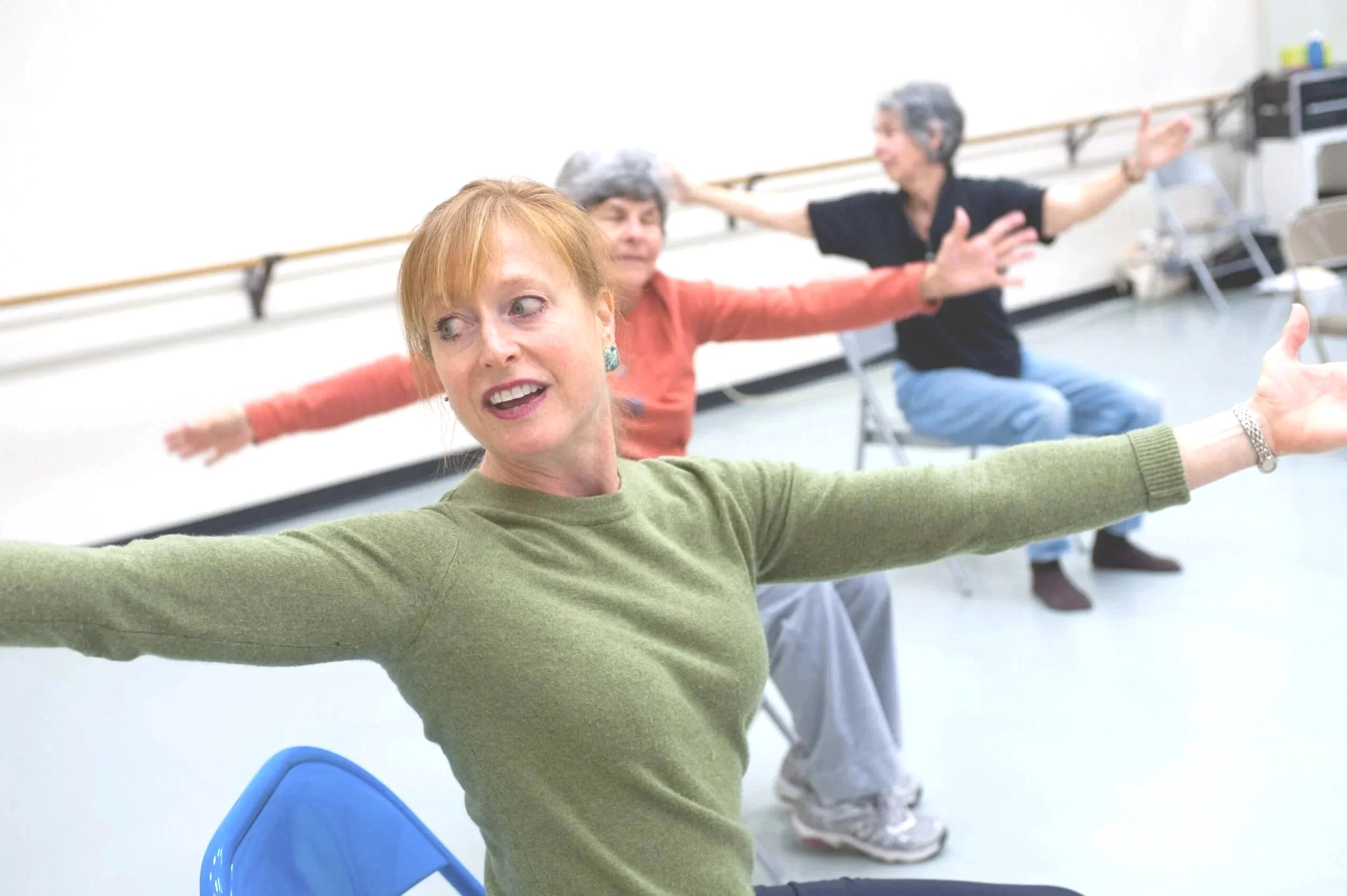Three women practicing ballet in a dance studio, seated on chairs with arms outstretched.