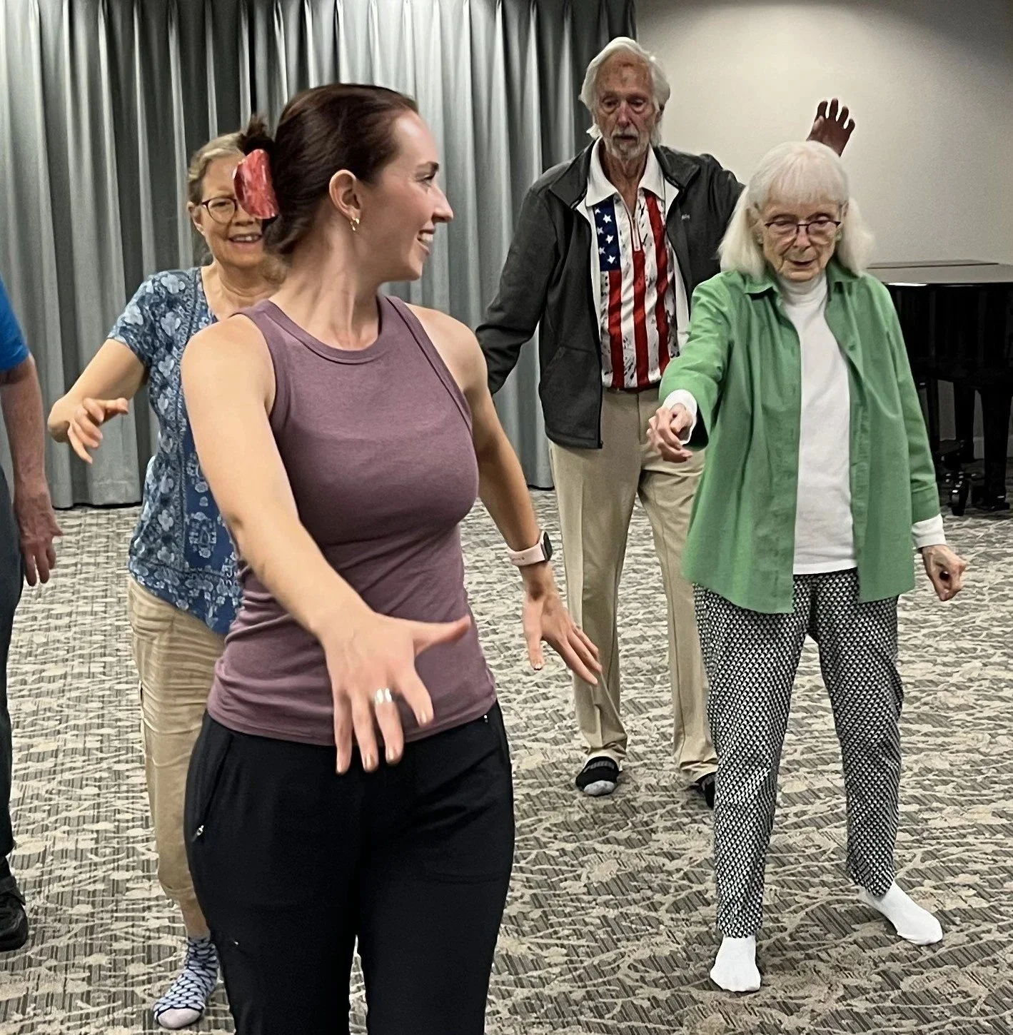Group of Dance for PD dancers dancing together in a room with a carpeted floor and gray curtains led by a dark-haired instructor, smiling and swinging her arms