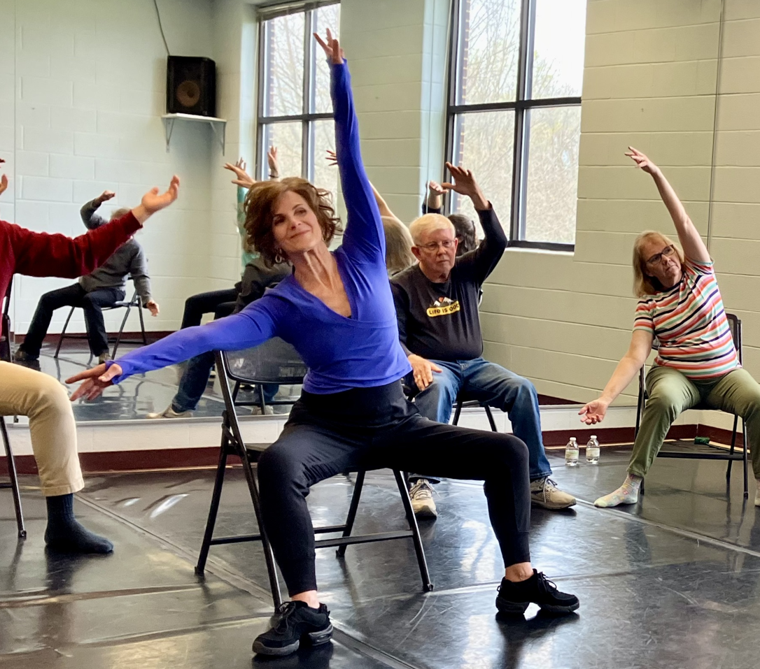 A group of people participating in a Dance for PD class, led by an instructor sitting on chairs and stretching with arms raised.
