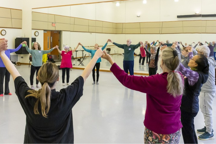 Group of adults holding hands in a circle during a dance class in a studio with mirrored walls.