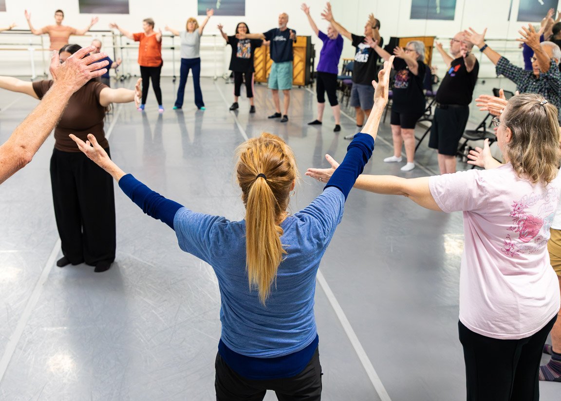 People participating in a Dance for PD class in a dance studio, standing in a circle with arms raised.