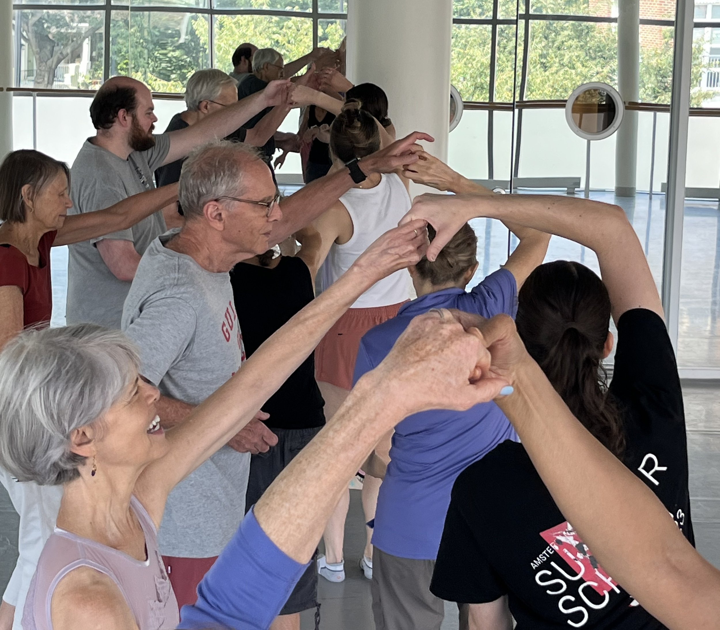 A group of people participating in a yoga or stretching class inside a bright room with large windows. The individuals are reaching forward or upward, focusing on their stretching poses.