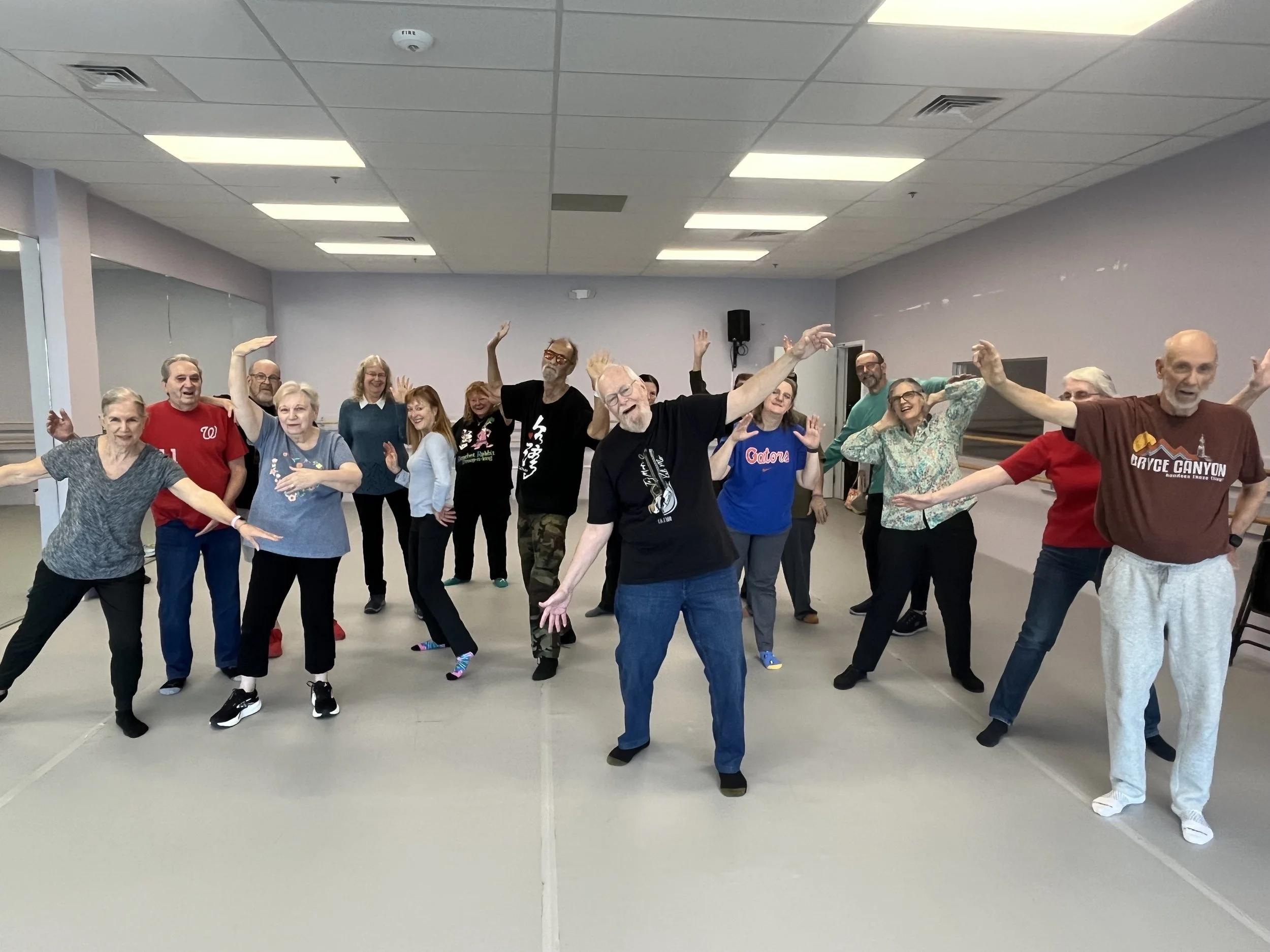 Group of Dance for PD participants posing for a photo, standing in various dance poses with arms outstretched or raised
