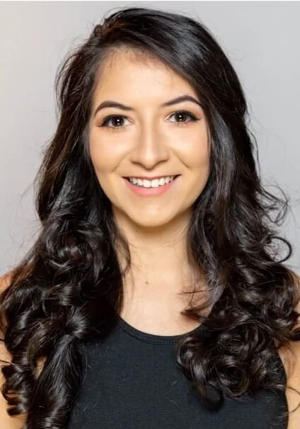 A young woman with long, dark, wavy hair smiling at the camera, wearing a black top.
