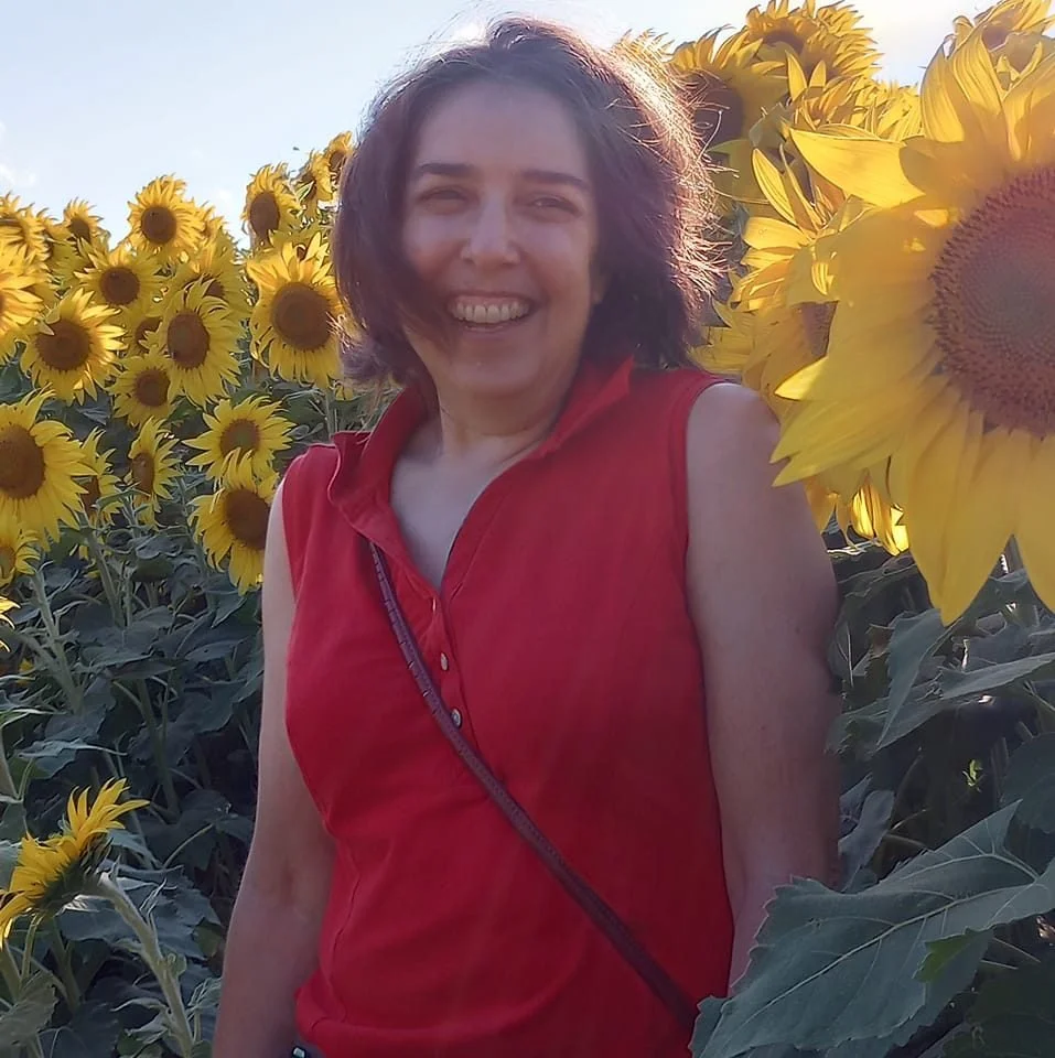 A woman smiling in a red sleeveless top standing in a sunflower field during daylight.