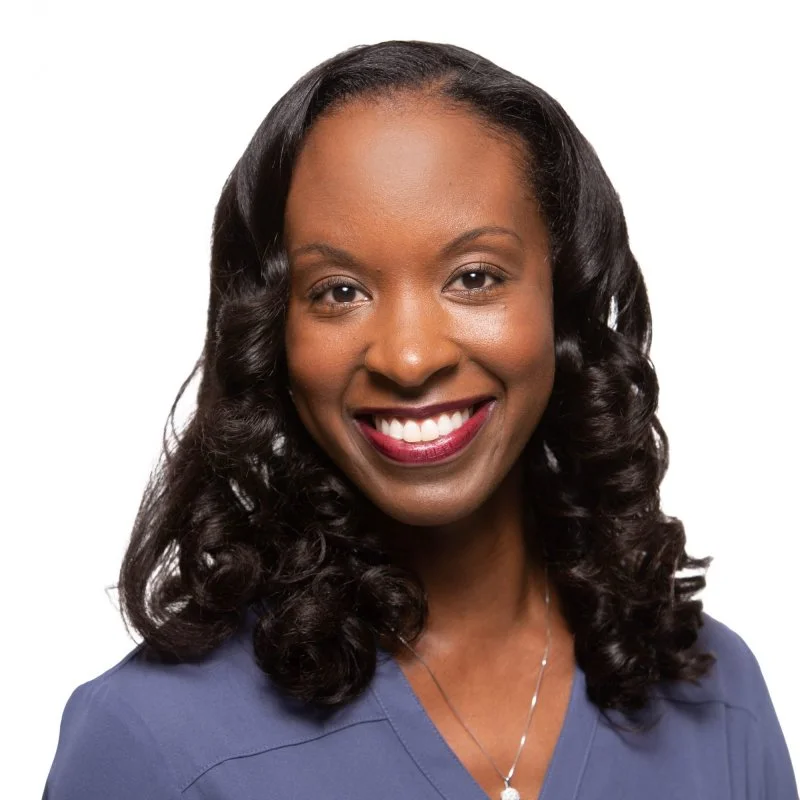 A woman with long, curly black hair smiling, wearing a blue top and a silver necklace.