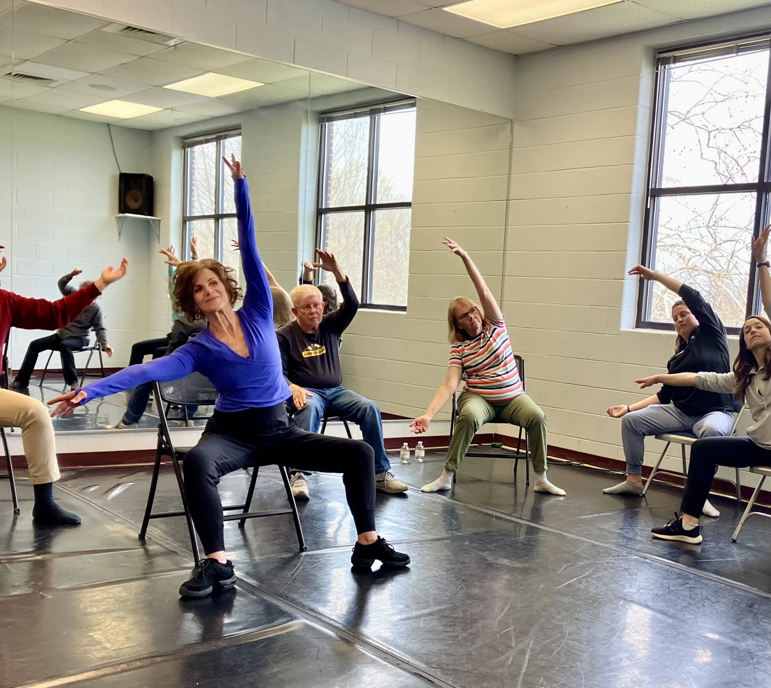 Group of people participating in a Dance for PD class in a dance studio, with large mirrors and windows in the background.