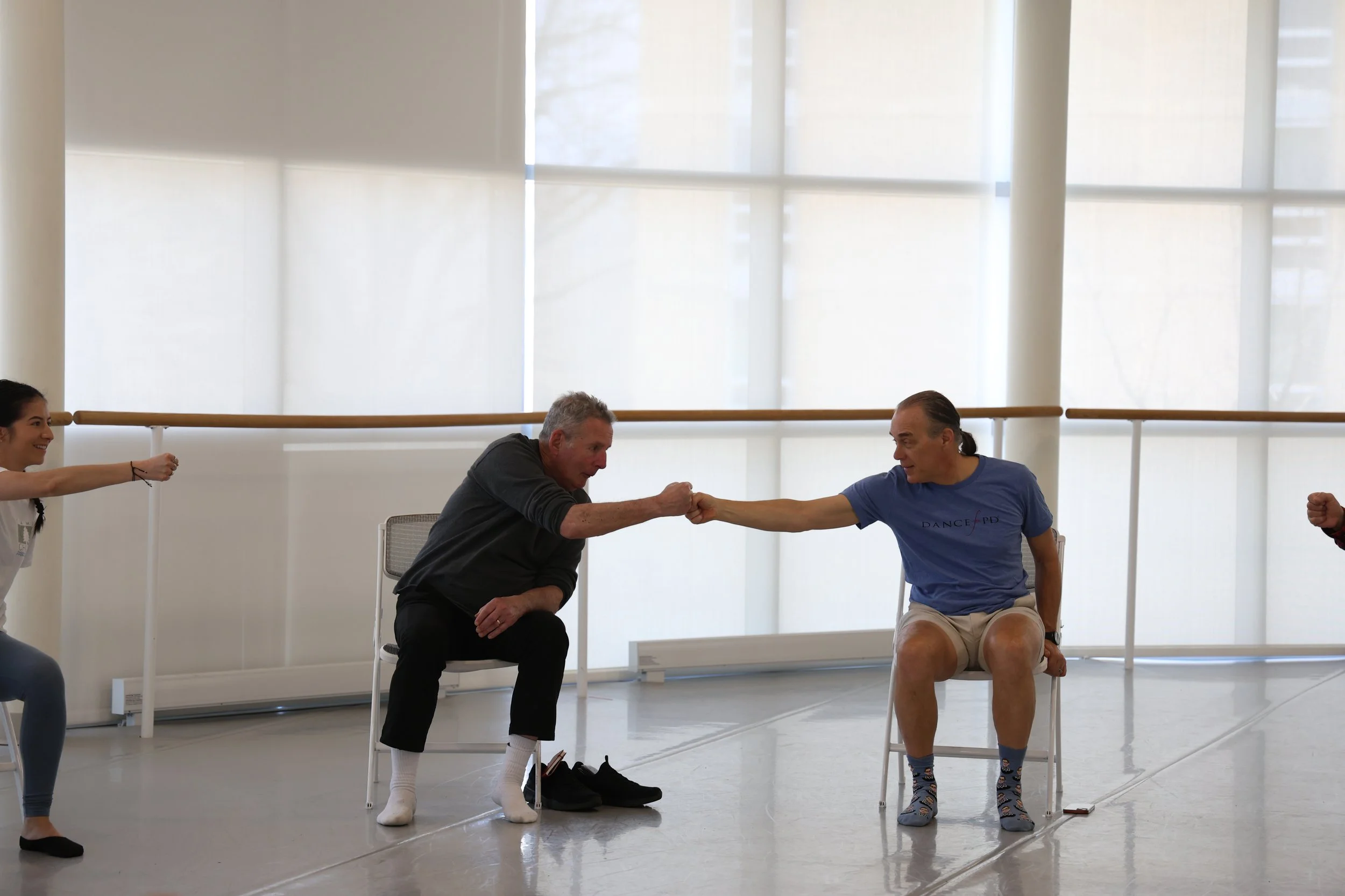 Two men sitting on chairs in a dance studio, reaching out and holding hands, with two women in the background, one on each side, stretching their arms forward.