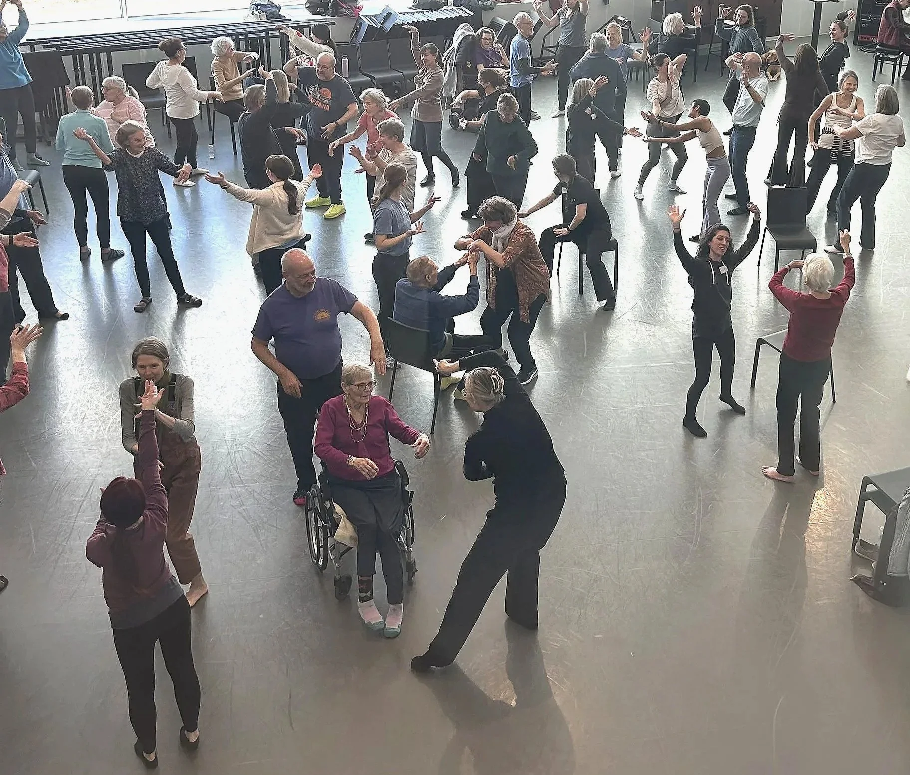 People dancing in a spacious hall, with some seated and others standing, including elderly individuals and individuals in wheelchairs.
