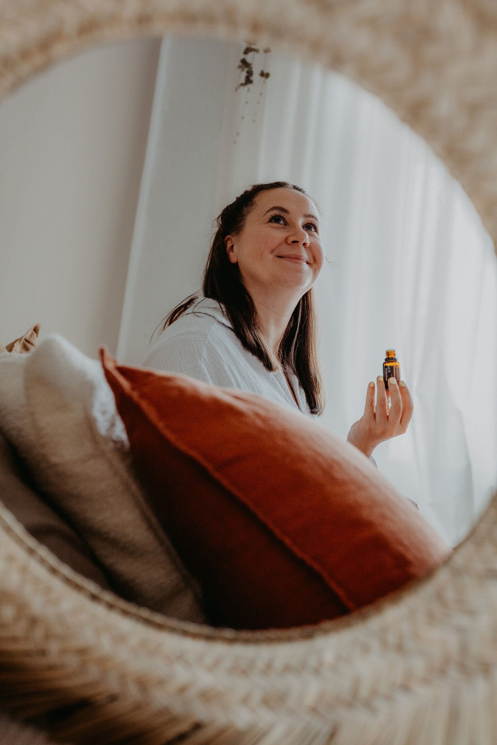 Frau schaut in einen Spiegel, hält eine Flasche ätherisches Öl, sitzt auf einem Sofa mit Kissen.