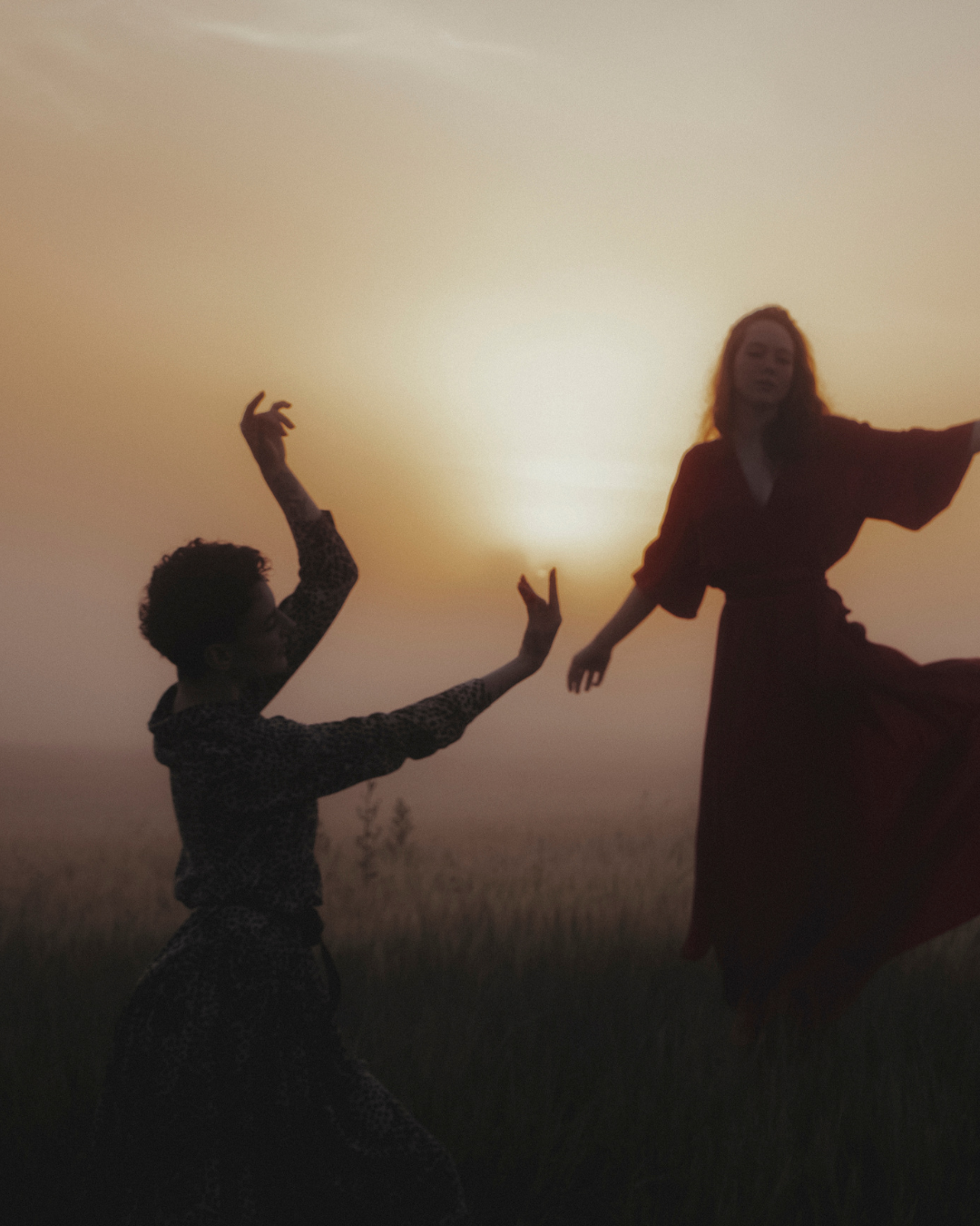 Zwei Menschen tanzen im Freien bei Sonnenuntergang, eine Person ist auf den Knien und streckt die Arme nach oben, die andere Person steht mit ausgestrecktem Arm.