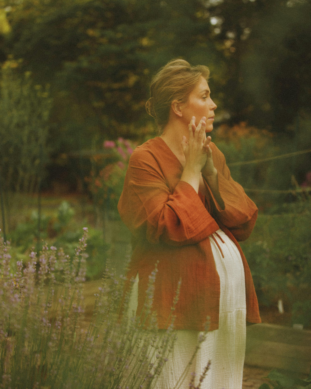 Eine schwangere Frau in einem Garten, die ihre Hände im Gesicht hält, im Profil sichtbar, mit grünen Bäumen und Blumen im Hintergrund.